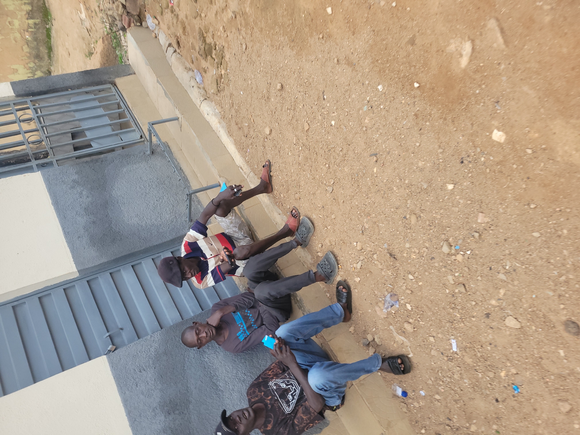 Three men are seated on the ground in front of a building. The building has a light-colored upper portion and a dark gray lower portion with a gray metal door and a gray metal gate. The ground is composed of dirt and gravel. The man on the far left is wearing a black baseball cap, a black t-shirt with a graphic print, and blue jeans. He is holding a blue object. The man in the middle is wearing a dark gray long-sleeved shirt and dark pants. He is resting his chin on his hand. The man on the far right is wearing a red, white, and blue striped t-shirt, dark pants, and a dark cap. He is holding a cell phone. All three men are wearing sandals. The scene appears to be outdoors, possibly in a semi-urban or rural area in Jos, Nigeria.