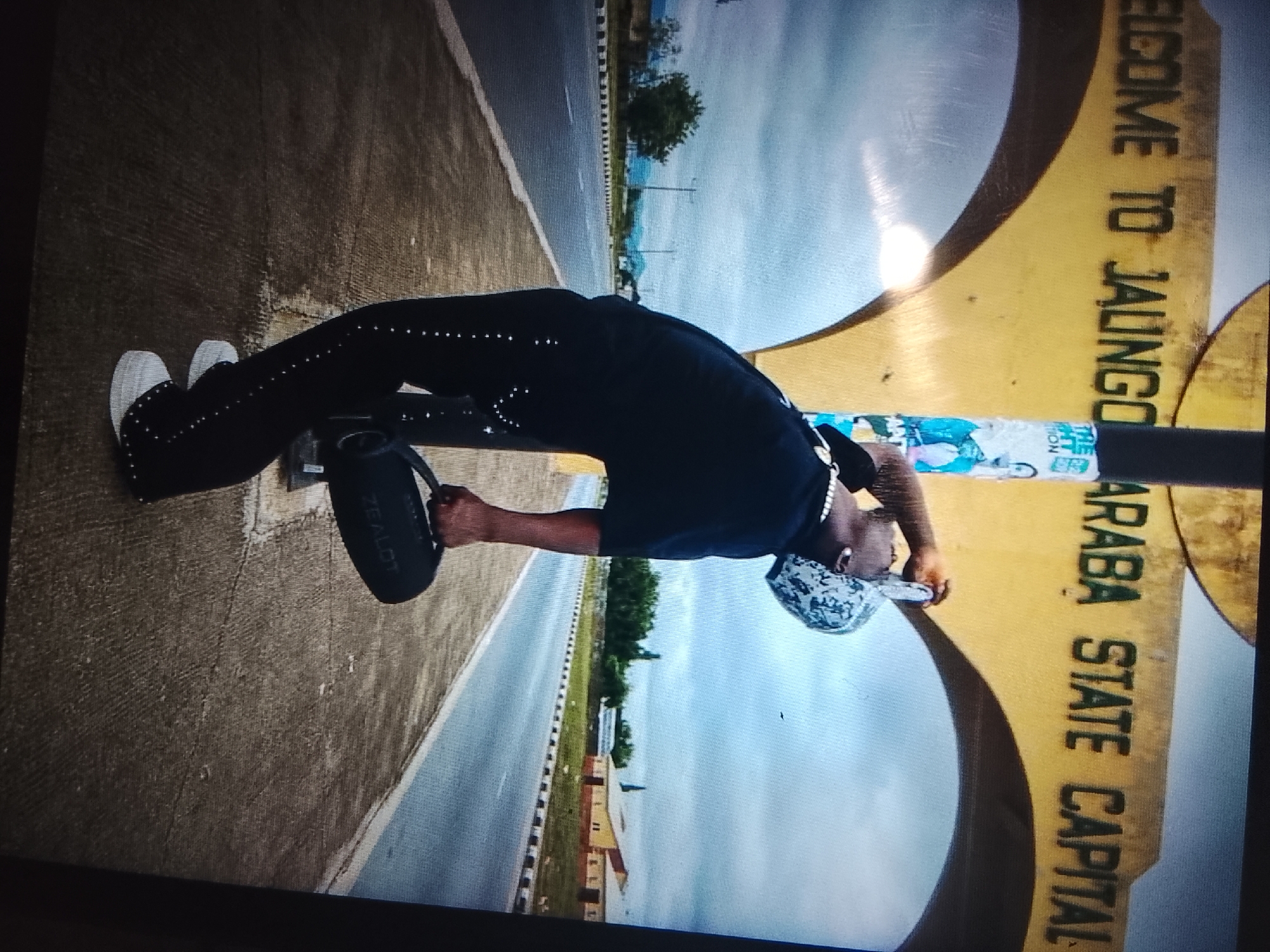 The image shows a person in black clothing and white shoes, holding a black cylindrical object. The person is positioned on a paved surface next to a road with a tree in the background. A large yellow sign with black text reads "WELCOME TO JALINGO ARABA STATE CAPITAL." The sky is cloudy. The scene appears to be an entrance to Jalingo, the capital city of Taraba State, Nigeria.