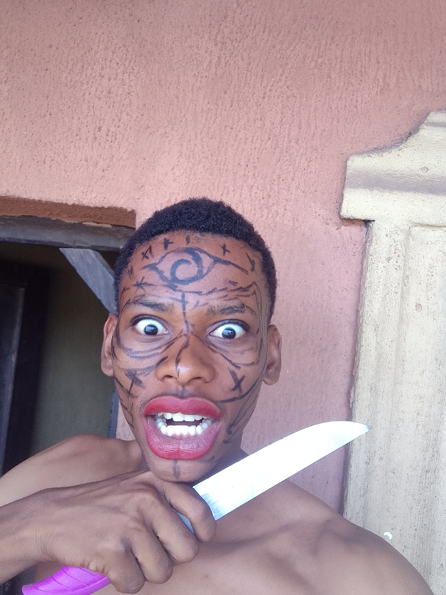 A close-up, eye-level shot captures a young Black person, likely male, from the chest up, against a textured, light reddish-brown wall. The person has short, dark, curly hair and is shirtless, revealing their shoulders and upper chest.
Their face is adorned with elaborate black markings resembling tribal or symbolic designs. A prominent eye-like symbol with a spiral is drawn on the forehead, and intricate lines and geometric shapes cover the cheeks, nose, and chin. Bright red lipstick is applied to their lips, which are wide open in an exaggerated expression, revealing white teeth. Their eyes are wide open, pupils dilated, conveying an intense or perhaps startled emotion.
The person holds a large kitchen knife with a shiny silver blade and a bright pink handle in their right hand. The knife is positioned in front of their chest, with the blade pointing upwards and slightly to the right.
The background consists of a textured wall, possibly stucco or rough plaster. To the left, a dark opening with a visible wooden frame suggests a doorway or window. To the right, a vertical section of a lighter, off-white or cream-colored textured wall or pillar is visible. The lighting appears to be natural and even, suggesting daytime. There is no visible text in the image.