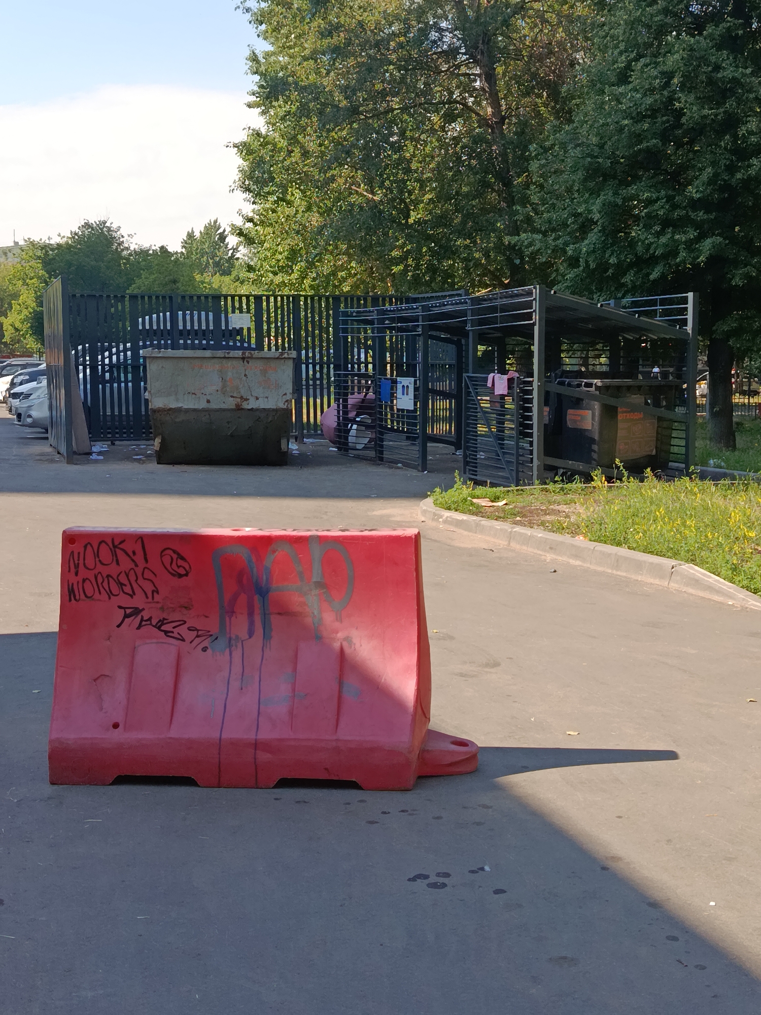 A red plastic water-filled barrier, spray-painted with graffiti, is positioned in the foreground on an asphalt surface. The graffiti on the barrier reads "NOOK 1 WONDERS" and "DAD". Behind the barrier, a black metal fence encloses a waste disposal area. Within the fenced area, a large, weathered metal dumpster is visible. To the right of the dumpster is a black metal structure with a roof, which appears to be a covered area for waste containers. In the background, trees and a bright blue sky with some clouds are present. This scene is located in Moscow, Russia, in the area of Troitsk, within the Troitsky Administrative Okrug.