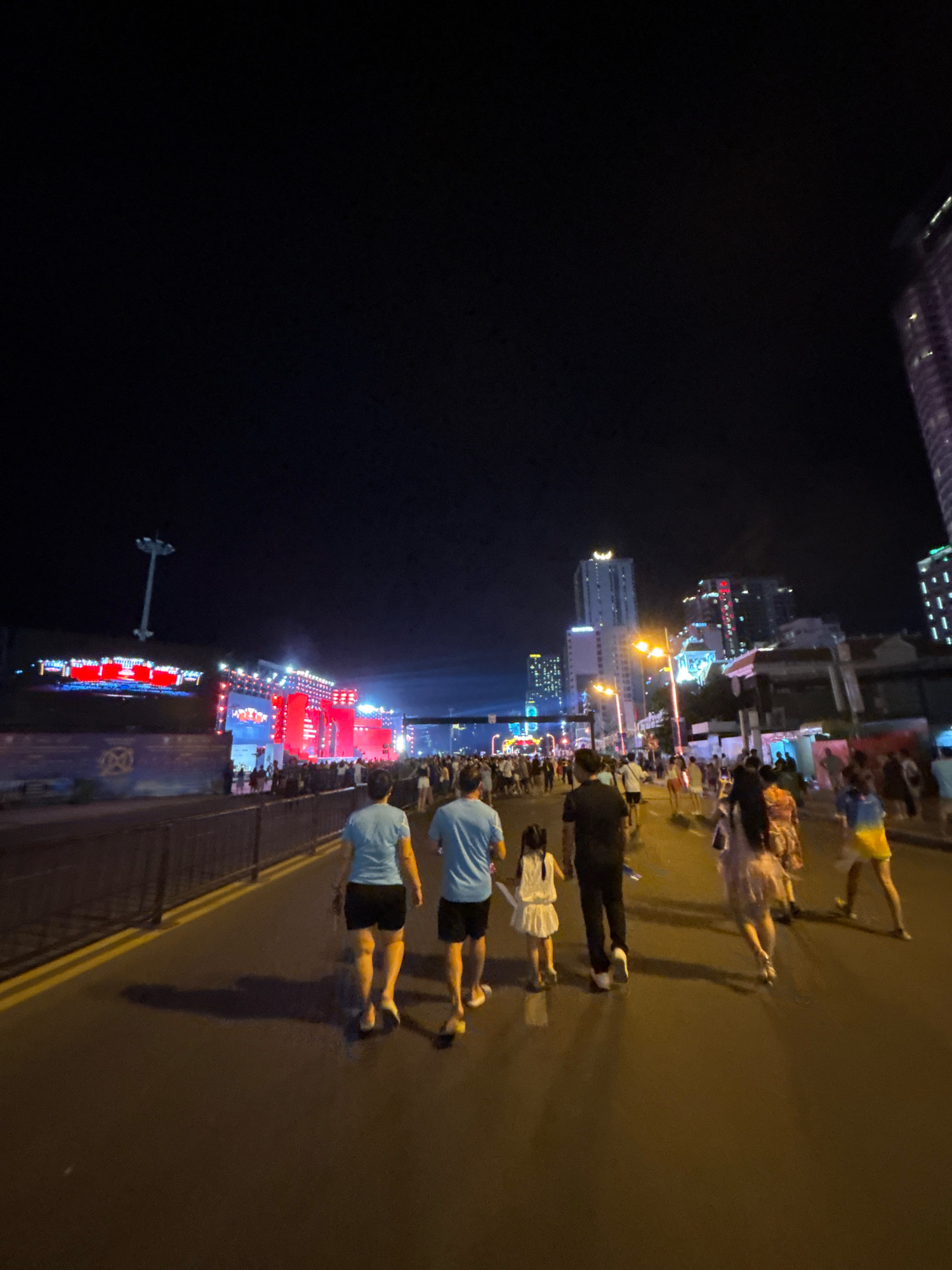 The image captures a night scene on a wide, paved walkway in Nha Trang, Vietnam, likely near the beachfront or a prominent entertainment area. A large, illuminated stage with bright red and blue lights is visible in the distance, suggesting a public event or concert. Numerous people are walking in both directions on the walkway, their figures silhouetted against the ambient light from the stage, streetlights, and surrounding buildings. In the foreground, two men in blue t-shirts and dark shorts walk away from the camera. To their right, a small girl in a white dress walks ahead of a man in black. Further down the walkway, other individuals and groups can be seen, including a woman in a patterned dress and a person in a yellow and blue outfit. Tall buildings with illuminated windows are visible in the background, indicating an urban setting. The dark night sky overhead contrasts with the brightly lit structures and the activity on the ground. The presence of yellow lines on the pavement suggests a designated pedestrian path or a roadway edge.