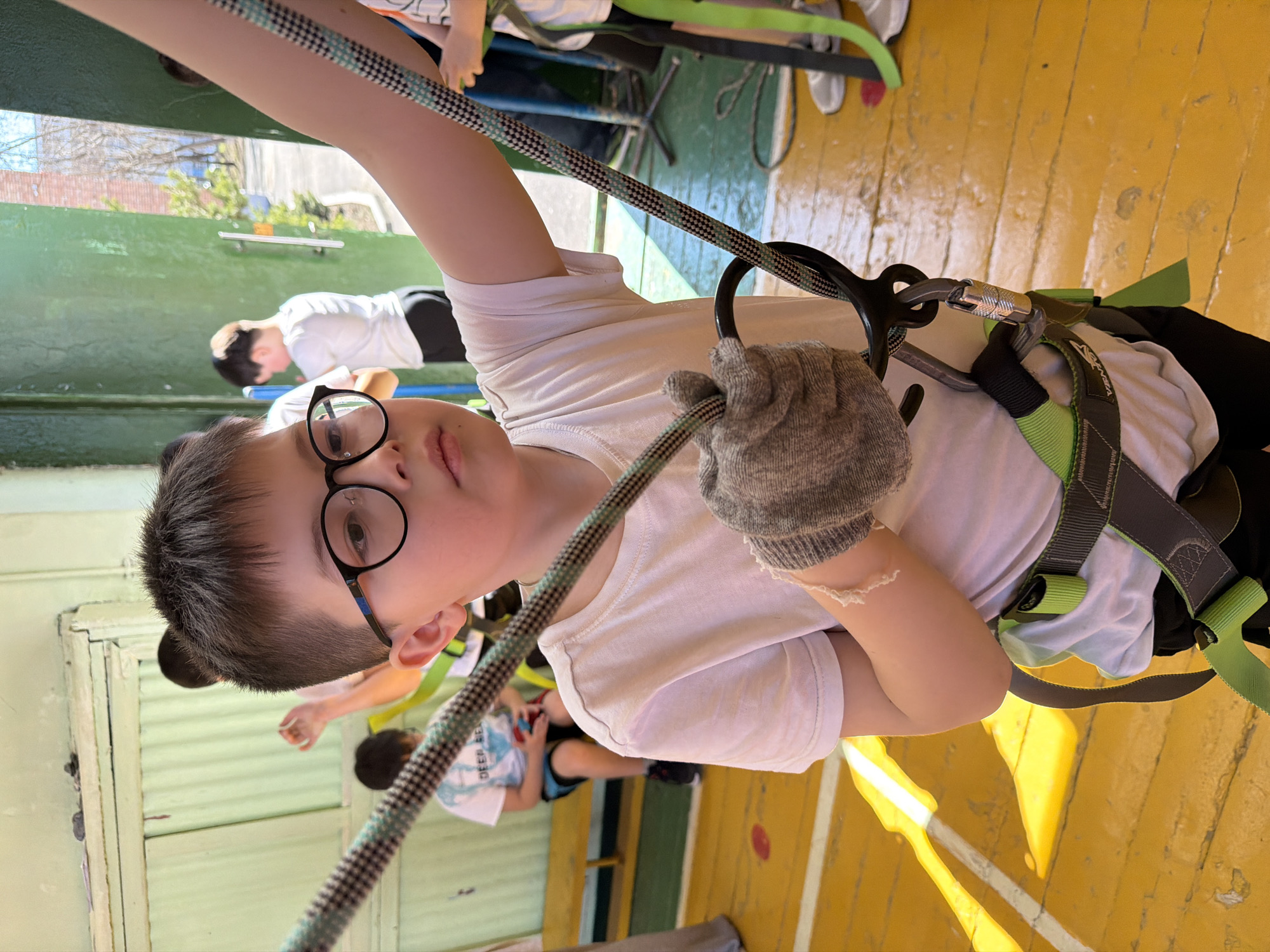A boy wearing glasses, a white t-shirt, and climbing gear stands indoors, holding a rope with a gloved hand. The location appears to be a training facility for climbing or ropes courses, with a painted yellow wooden floor. Other people are visible in the background, also possibly participating in climbing-related activities. The image was taken at an angle, giving a rotated perspective of the scene. The location is the city of Izhevsk, Russia.