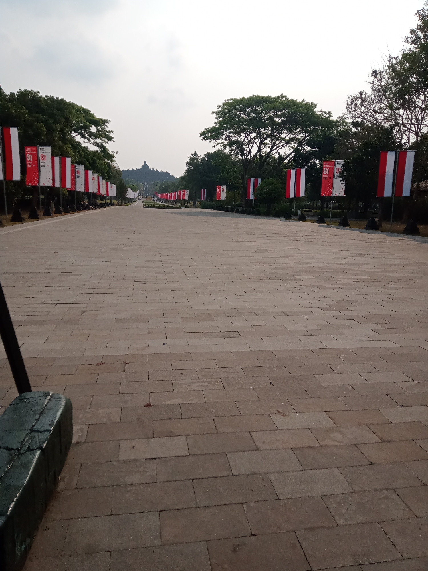 The image shows a wide, paved pathway leading towards a distant structure that appears to be a temple or monument, possibly Borobudur Temple in Indonesia, given its distinct silhouette against the horizon. The pathway is lined on both sides with tall, vertical banners. These banners alternate between being predominantly white with red stripes on the sides and red with white stripes on the sides. Many of the red banners also feature the number "81" in white, suggesting an anniversary or commemoration.

The pathway itself is constructed from large, rectangular stone tiles laid in a stretcher bond pattern. The scale of the pathway indicates it is a ceremonial or significant thoroughfare. On the left side of the foreground, a dark, weathered stone object, possibly a bench or a planter base, is partially visible, providing a sense of scale and indicating the material used in the surrounding landscaping.

The environment is characterized by a bright, overcast sky, casting a diffused light over the scene. Lush green trees with full canopies flank the pathway on both sides, creating a natural border and framing the view towards the distant monument. The trees on the left are denser, with broad leaves, while those on the right are more varied in type and density. The overall impression is one of an open, grand space, likely a tourist destination or a significant public area within the city of Borobudur, Indonesia, decorated for a celebratory event.