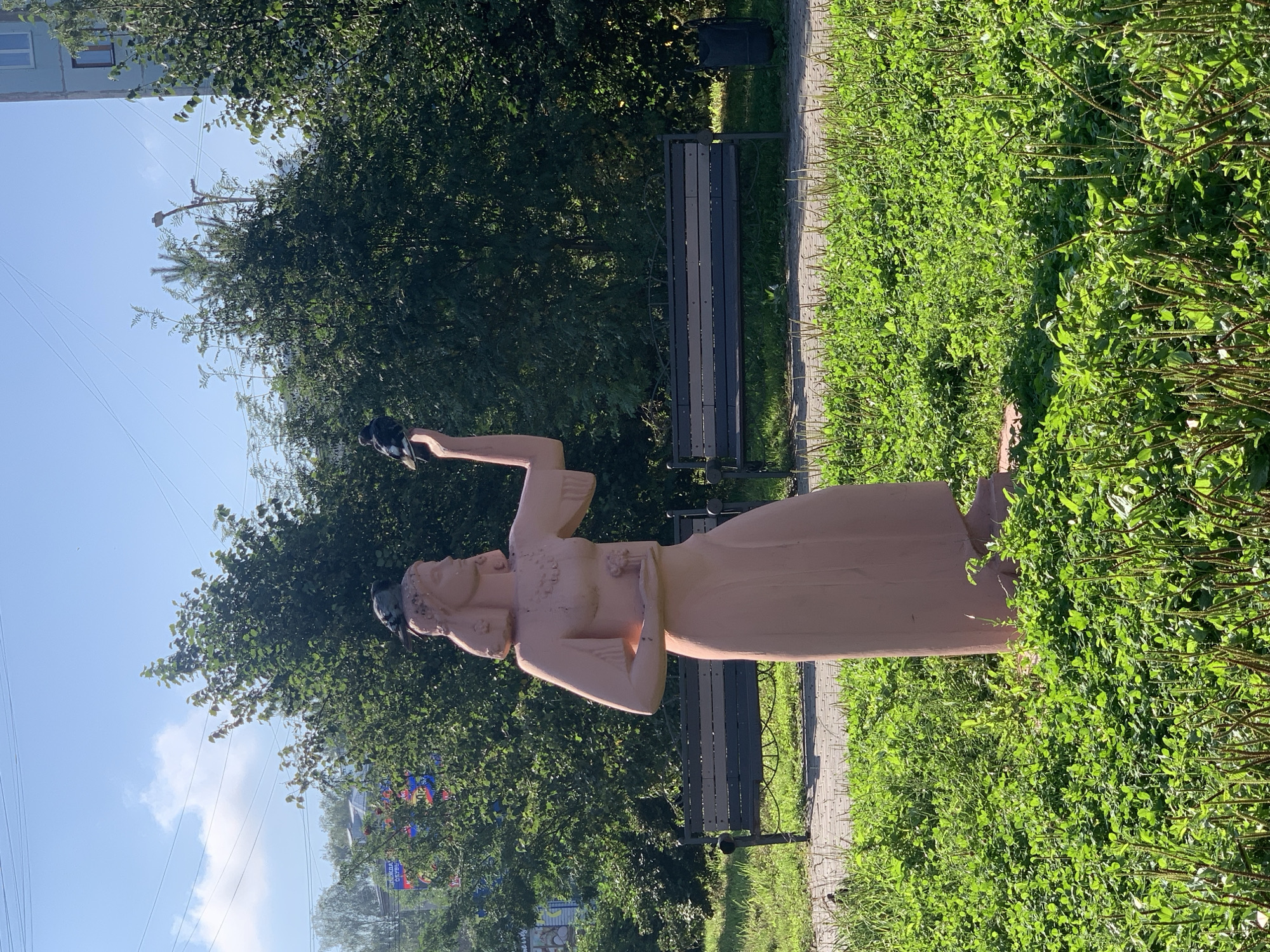 A full-shot photograph shows a light pink, weathered statue of a woman standing in a park in Prokopyevsk, Russia. The statue depicts a woman with her arms extended, holding a flower in her left hand and reaching upwards with her right. Three pigeons are perched on the statue: one on her head, one on her extended hand, and one on her left shoulder. The statue stands in a patch of lush green grass with some sparse weeds and clover. In the background, two dark-colored park benches with metal frames are visible, along with a black trash can. Behind the benches, a dense line of green trees forms a natural backdrop, with a blue sky and scattered white clouds overhead. The overall impression is that of a serene, sunlit park scene.
