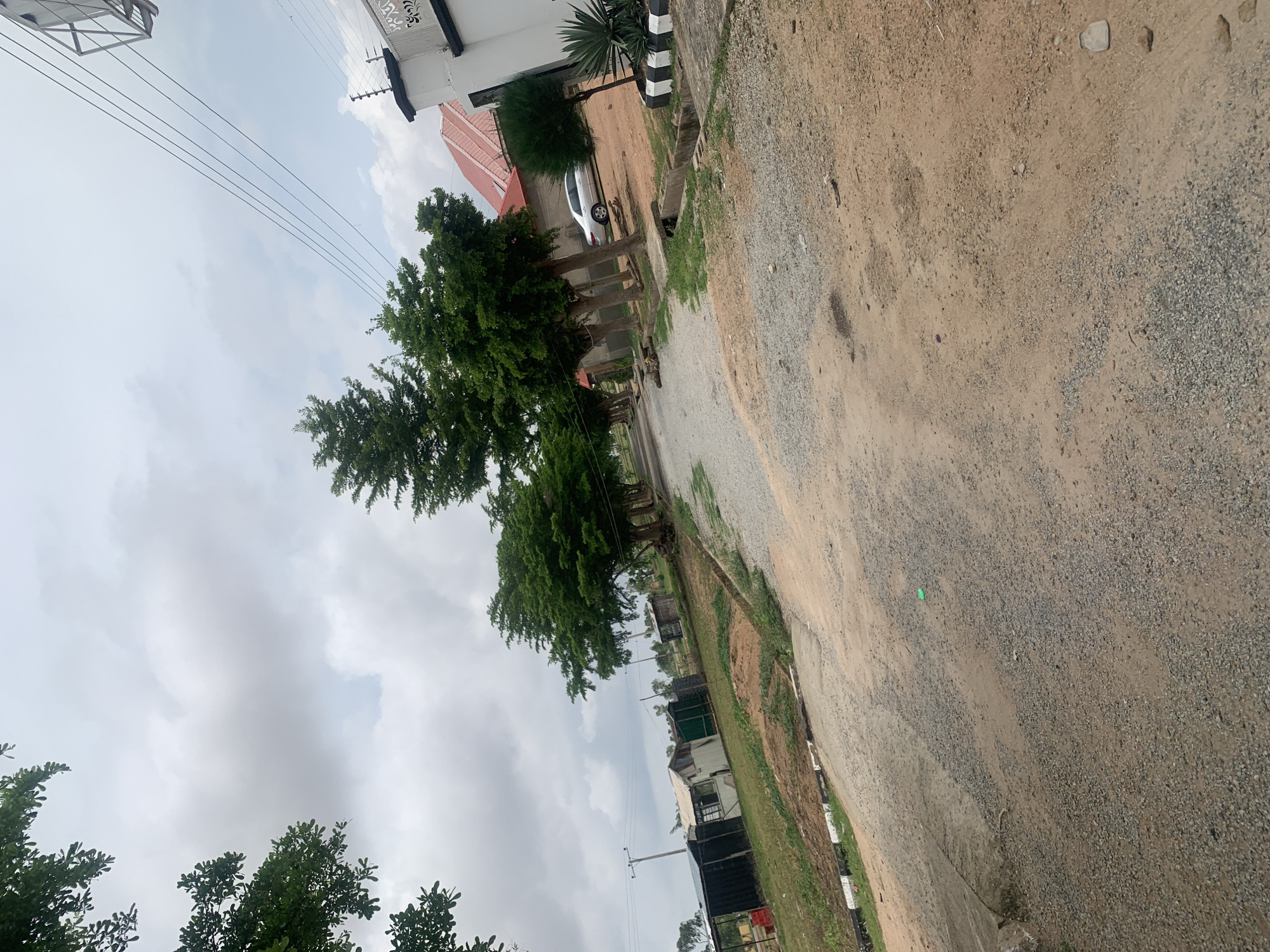 The image shows a dirt road in Jauro Yinu, Nigeria. On the left side of the road are several small, makeshift structures, some appearing to be stalls or shelters. Beyond these structures, there are trees and grassy areas. A power line runs across the upper part of the image. On the right side of the road, there is a white building with a red-tiled roof, an electric fence on top of the wall, and a silver car parked in front of it. The road itself is unpaved and covered with gravel and dirt. There are trees lining the right side of the road, and a concrete drainage ditch runs alongside the road. The sky is cloudy and overcast.