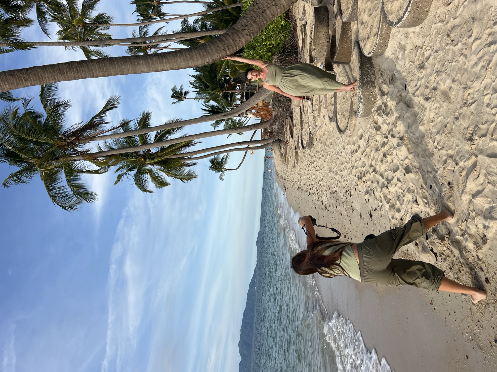 This media file depicts a bright, sunny day on a tropical beach, likely in Baan Tai, Thailand. The scene is dominated by tall palm trees with lush green fronds, framing a clear blue sky with scattered wispy clouds. The ground is covered in soft, light-colored sand, with footprints visible in some areas.
Two individuals are present. In the foreground, a person with long brown hair is lying on the sand, holding a black camera and appearing to take a photograph. They are wearing a light green tank top and loose, olive-green capri pants. Their body is positioned towards the ocean, suggesting they are capturing a view of the water or the landscape beyond.
Further back, another person is standing, leaning against the trunk of a large palm tree. They are wearing a flowing olive-green dress and appear to be posing or looking towards the camera. Their arm is extended upwards, touching the tree trunk, and they are smiling. The beach extends into the background, with the gentle waves of the ocean lapping at the shore. In the distance, the silhouette of a mountainous landscape can be seen against the horizon.
The lighting suggests it is daytime, likely morning or afternoon, with the sun casting shadows on the sand and highlighting the textures of the beach and the palm trees. The overall atmosphere appears relaxed and serene, with the natural beauty of the tropical environment being the central focus. There is no visible text in the image.