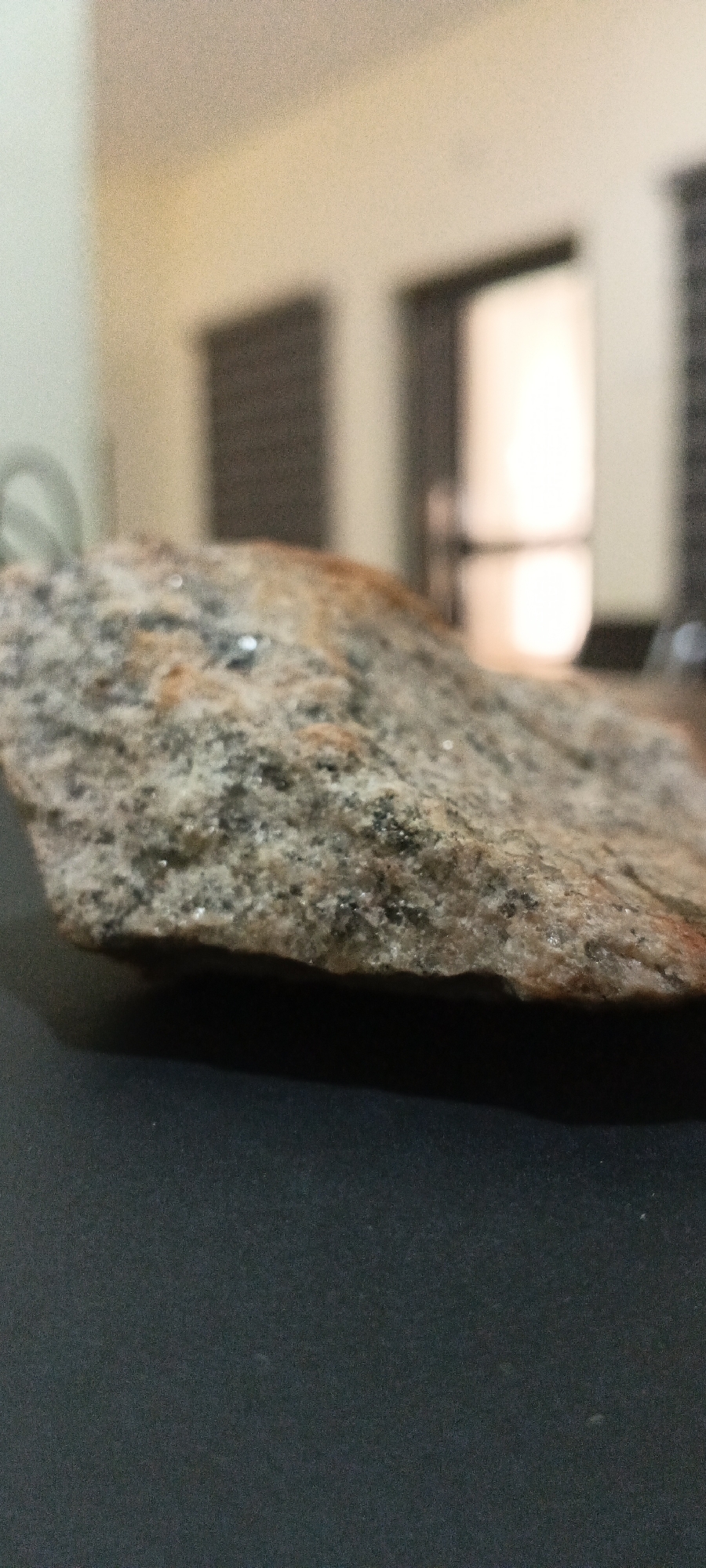 A close-up, shallow depth-of-field photograph focuses on a rough, granular rock resting on a dark, flat surface. The rock occupies the lower-middle portion of the frame, extending from the left edge towards the right. Its surface is textured with visible mineral grains, displaying a mix of muted gray, black, white, and reddish-brown tones. Numerous small, reflective specks, possibly mica or other metallic minerals, are scattered across its surface, creating highlights. A particularly bright, circular reflection is prominent on the upper surface of the rock, suggesting a direct light source above it. The rock casts a soft shadow on the dark surface beneath it.
The background is heavily blurred and out of focus, indicating an indoor setting. It features a light-colored wall on the left, transitioning to a warmer, yellowish-beige tone towards the center-right. Distant, dark, rectangular shapes, possibly window frames or blinds, are vaguely visible. A very bright, vertically elongated light source, likely a window or door, is blurred in the background, creating the prominent reflection on the rock. There is no discernible activity, people, or visible text in the image. The lighting appears to be either natural light from a window or strong artificial light. While the location context "Shaba, Nigeria" is provided, the image itself does not contain specific visual cues to confirm this geographical location.