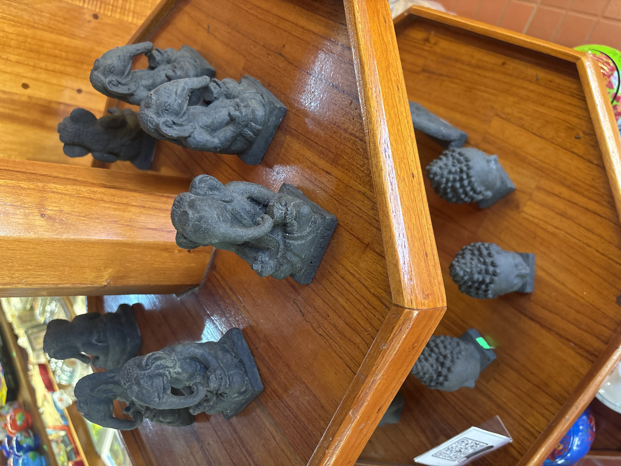 The image shows a display shelf in what appears to be a souvenir shop. The shelf is made of polished wood and is divided into two tiers. On the top tier, several small, dark gray elephant figurines are arranged in a semi-circle. These figurines appear to be made of stone or resin and are detailed with trunks, ears, and legs. In the background, out of focus, are other items for sale, including what looks like colorful ceramic teapots. On the lower tier, there are three small, dark gray Buddha head figurines with curly hair, also arranged on a wooden shelf. A small white stand with a QR code is visible on the left side of the lower shelf. The overall impression is of a retail display of religious or cultural souvenirs. The reference to Đà Nẵng, Vietnam, suggests this might be a shop in that city.
