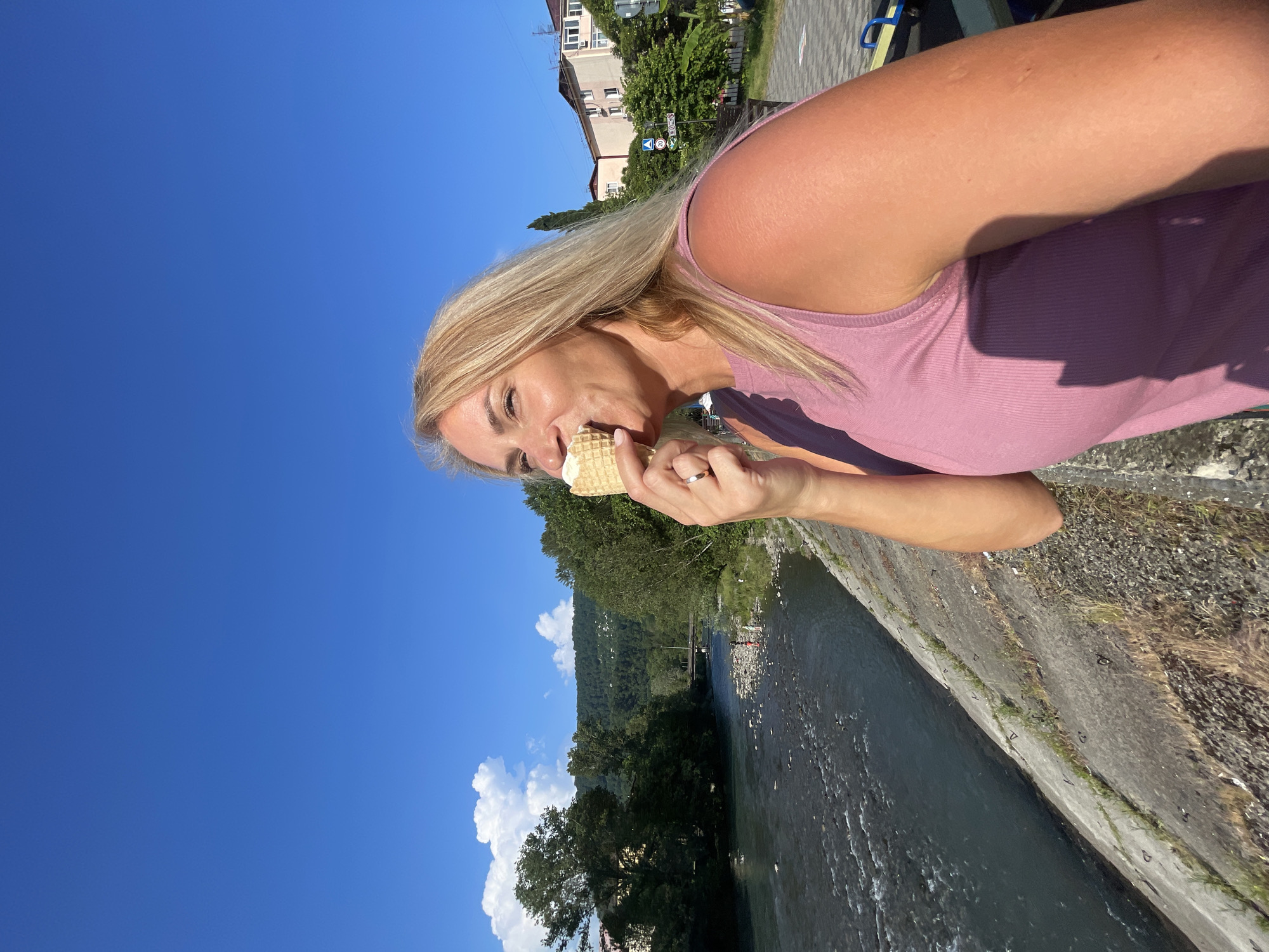 A blonde woman with fair skin is shown in a medium shot, holding an ice cream cone to her mouth and eating it. She is wearing a pink sleeveless top and a gold wedding band on her left hand. The woman is positioned in the foreground, with her face turned towards the right, looking upwards.
Behind her, a river flows with visible ripples. The riverbank is lined with a concrete embankment with some greenery growing through it. In the background, lush green trees and hills are visible, suggesting a natural landscape. Further in the distance, buildings and a street can be seen. The sky is a clear, vibrant blue with a few white clouds.
The scene appears to be in Sochi, Russia, based on the presence of buildings with architectural styles common in the region and the natural environment. The lighting suggests it is daytime, likely a sunny afternoon given the shadows and the brightness of the sky.