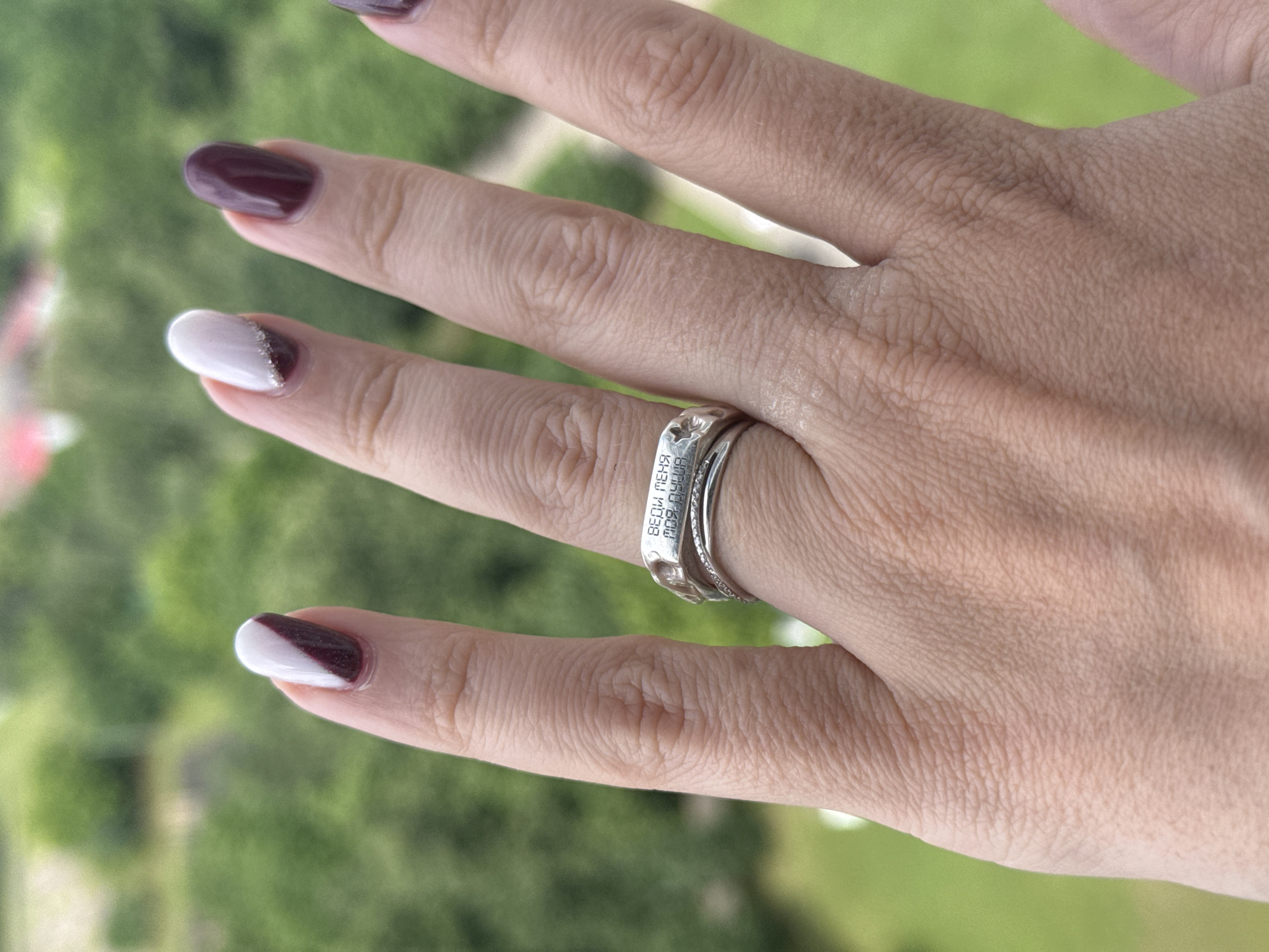 An overhead medium shot shows a person's left hand with a ring on the ring finger, against a blurred background of greenery and some buildings, suggesting an outdoor setting, possibly in Moscow, Russia. The hand has a light skin tone and well-manicured nails painted in a two-tone design of white and deep red, with some subtle glitter on the ring finger's white portion. The ring on the ring finger is a silver band with an inscription on its surface, which appears to be in Cyrillic. The inscription is partially visible and reads "ВЕДИ МЕНЯ" which translates to "LEAD ME" and "МОЯ ДУША" which translates to "MY SOUL". The ring is stacked with another thin, delicate silver band. The image is sharp on the hand and ring, with the background out of focus.