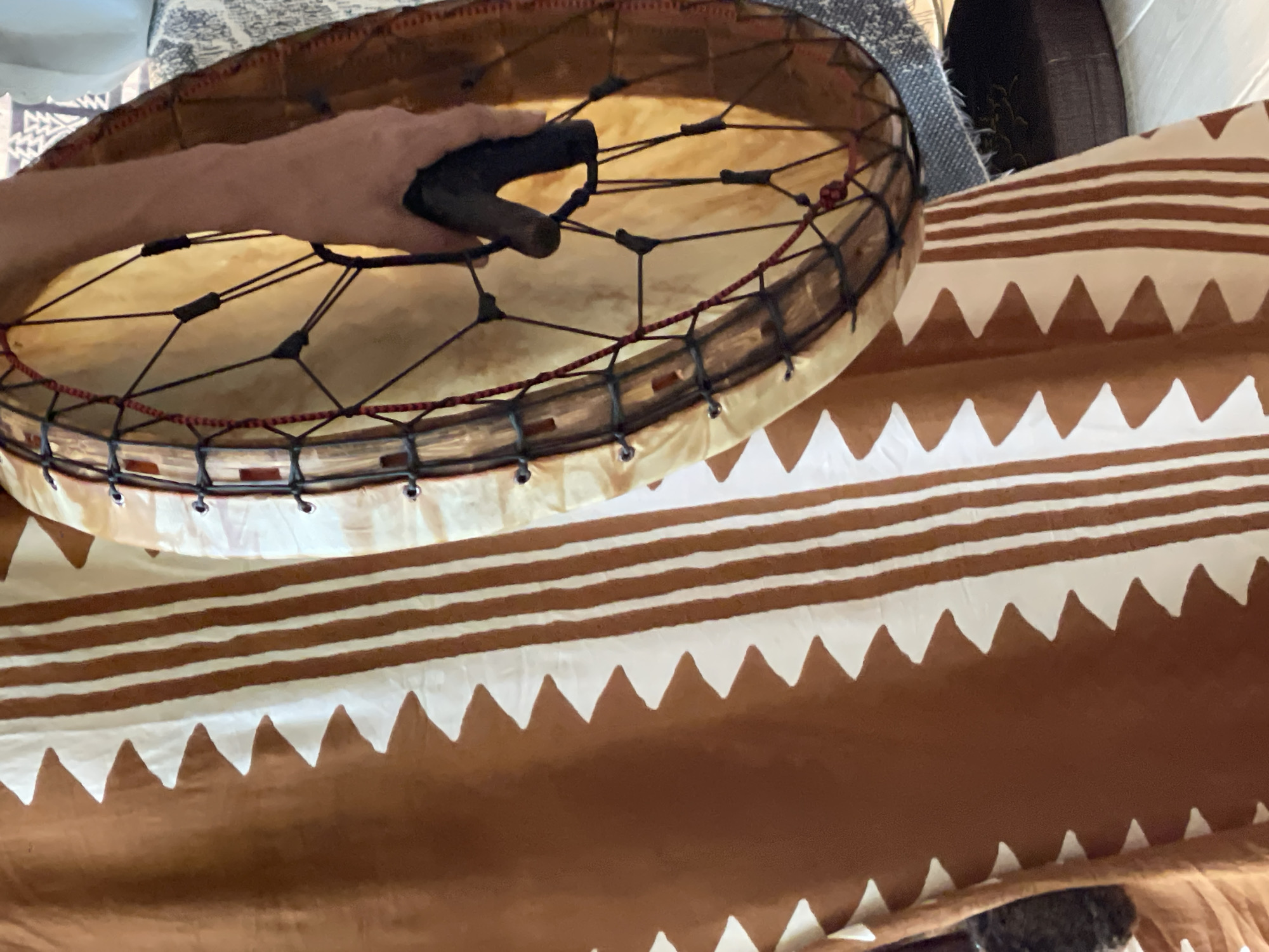 The image shows a close-up view of a person holding a drum. The person is wearing a long, brown and white patterned robe. The robe features a zigzag pattern on the left side and vertical stripes on the right. The drum is circular with a light-colored skin and is held by a hand grasping a dark mallet. The drum is laced with black cord and accented with red string. The background shows a textured surface, possibly a rug or blanket, and a dark circular object. The lighting suggests an indoor setting. The location is not specified.