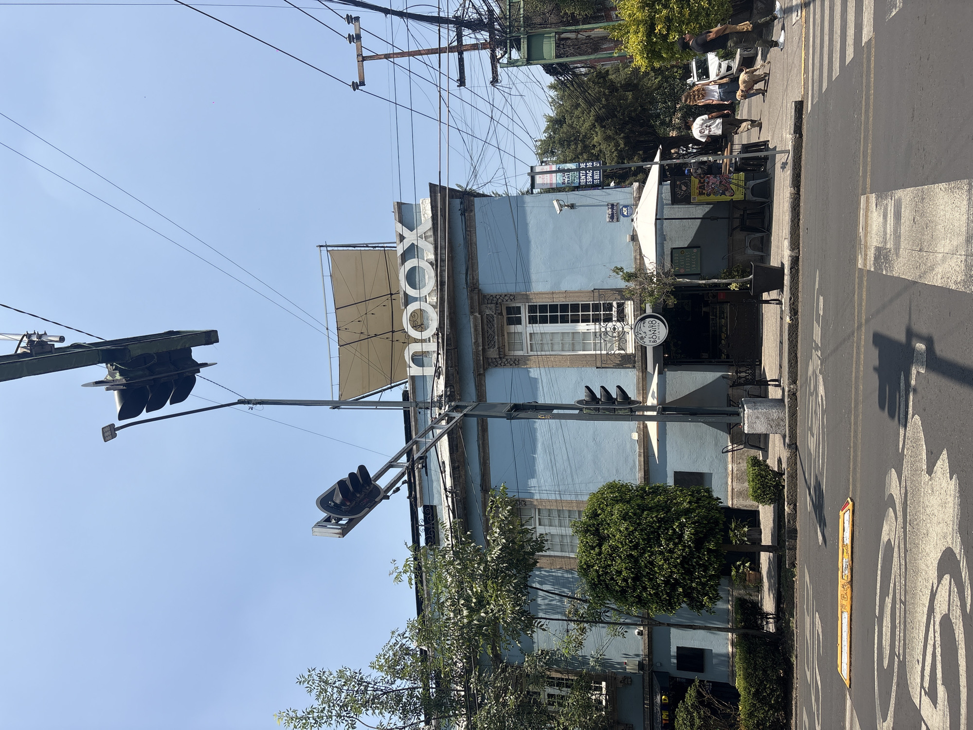 The image captures a street scene in Mexico City, Mexico, specifically at the intersection of Calle de Querétaro and another street. The building on the corner is painted light blue and has "NOOX" in large white letters on its roofline, possibly the name of a business located within the building. A sign on the building lists "CAFE BEBIDAS SNACKS" and another says "BONITO CAFE". The building features white-framed windows and a white awning extending over its entrance.
A traffic light standard is prominently visible on the left side of the frame, with two sets of traffic lights, one aimed at traffic traveling left to right and another aimed right to left. Numerous overhead utility lines crisscross the sky.
On the right side of the image, there is a crosswalk with pedestrians, including at least two people walking a dog. The street itself is marked with road lines. There are also other people visible. The environment suggests a sunny day.