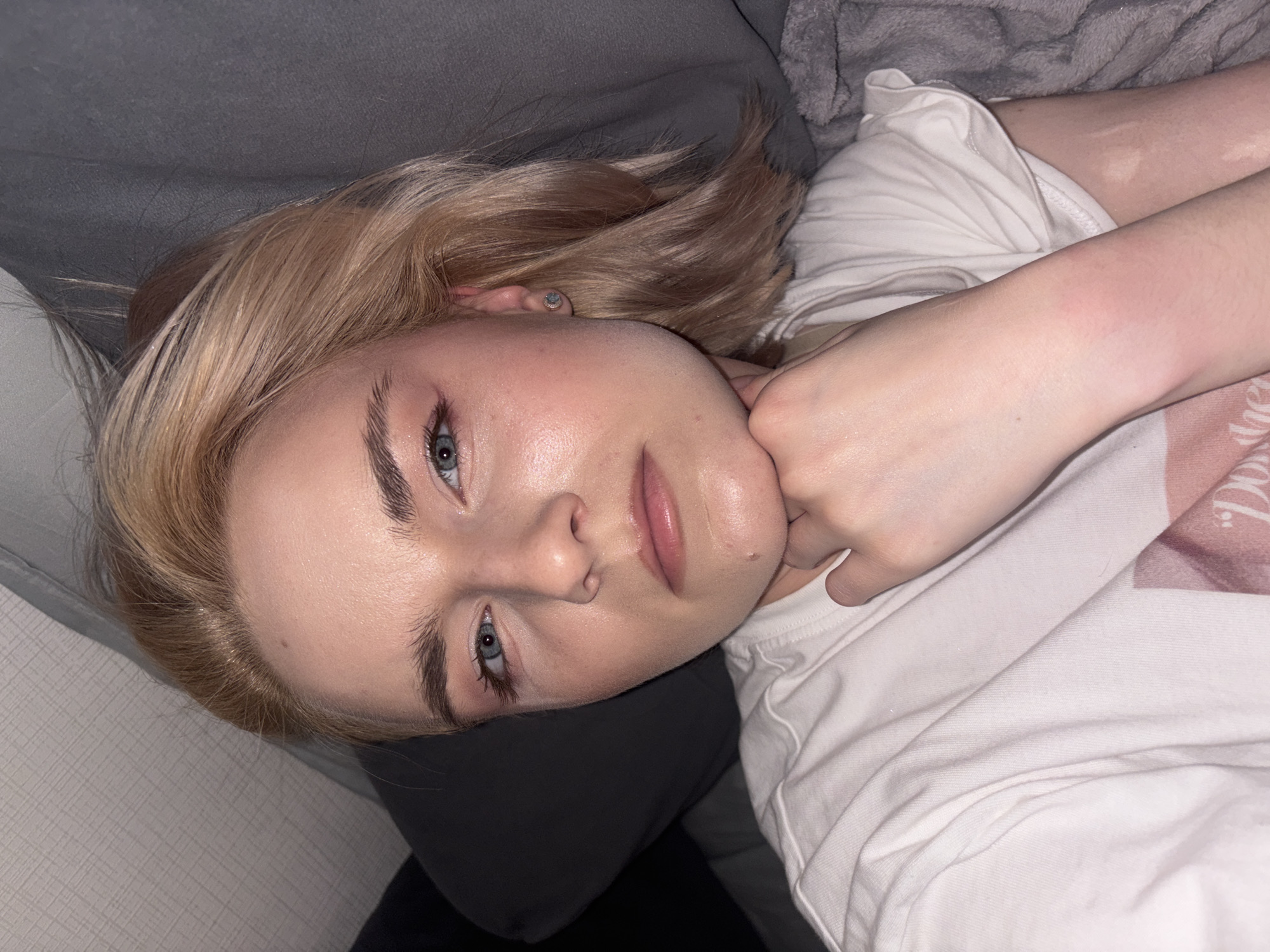 A young person with fair skin, light blonde hair, and blue eyes is depicted lying on their left side, looking directly into the camera. The image is rotated 90 degrees counter-clockwise. The individual's right hand is positioned under their chin/jawline. Their eyes feature visible dark eyeliner and mascara, complementing defined dark eyebrows. Their lips are light pink and slightly parted. A small mole is present on the right side of the chin. The right ear is partially visible, adorned with a small silver stud earring. The person is wearing a white t-shirt, with a faint pink graphic partially visible on the right chest area. They are resting on bedding that includes dark gray fabric on the left side and a mix of white and light gray, textured fuzzy fabric on the right side of the frame. The lighting appears soft and indirect.
