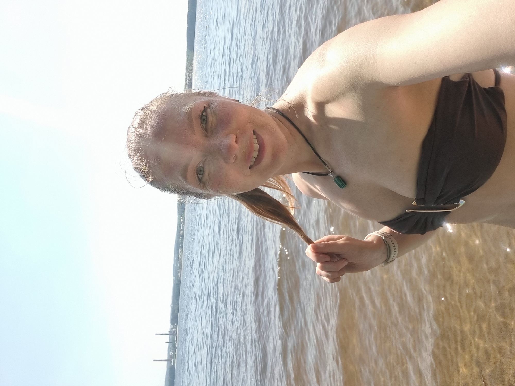 A woman in a black bikini stands in shallow water, holding a strand of her blonde hair. She is smiling at the camera. She wears a dark necklace with a green pendant and a smartwatch on her left wrist. The background features a body of water under a clear sky, with distant buildings or structures along the shoreline. The location is identified as Chaykovsky, Russia.