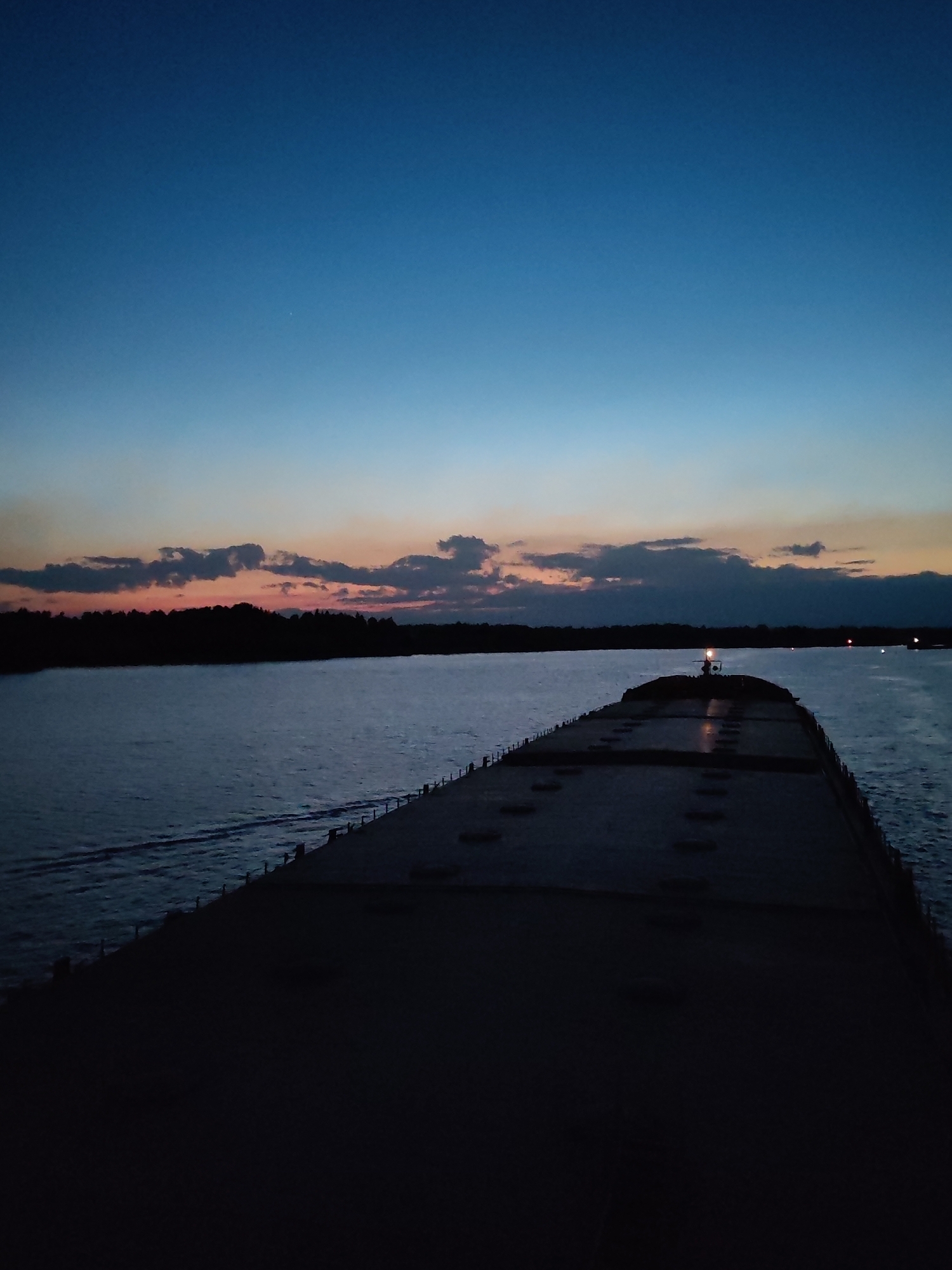 A low-angle photograph captures the aft section of a barge during twilight. The barge, constructed with a flat top, dominates the foreground, the deck appearing a dark silhouette. The edges of the barge are outlined by small, uniformly spaced vertical structures, possibly stanchions or railings.
Beyond the barge, a body of water reflects the colors of the sunset, with tones of orange and pink appearing through the clouds. The opposite shore is visible, lined with trees silhouetted against the fading light. Two small lights are seen on the water in the distance.
The sky is gradient of deep blue, transitioning into lighter shades towards the horizon where the sunset colors are most apparent.
There are no apparent people in the image. The location is not indicated.