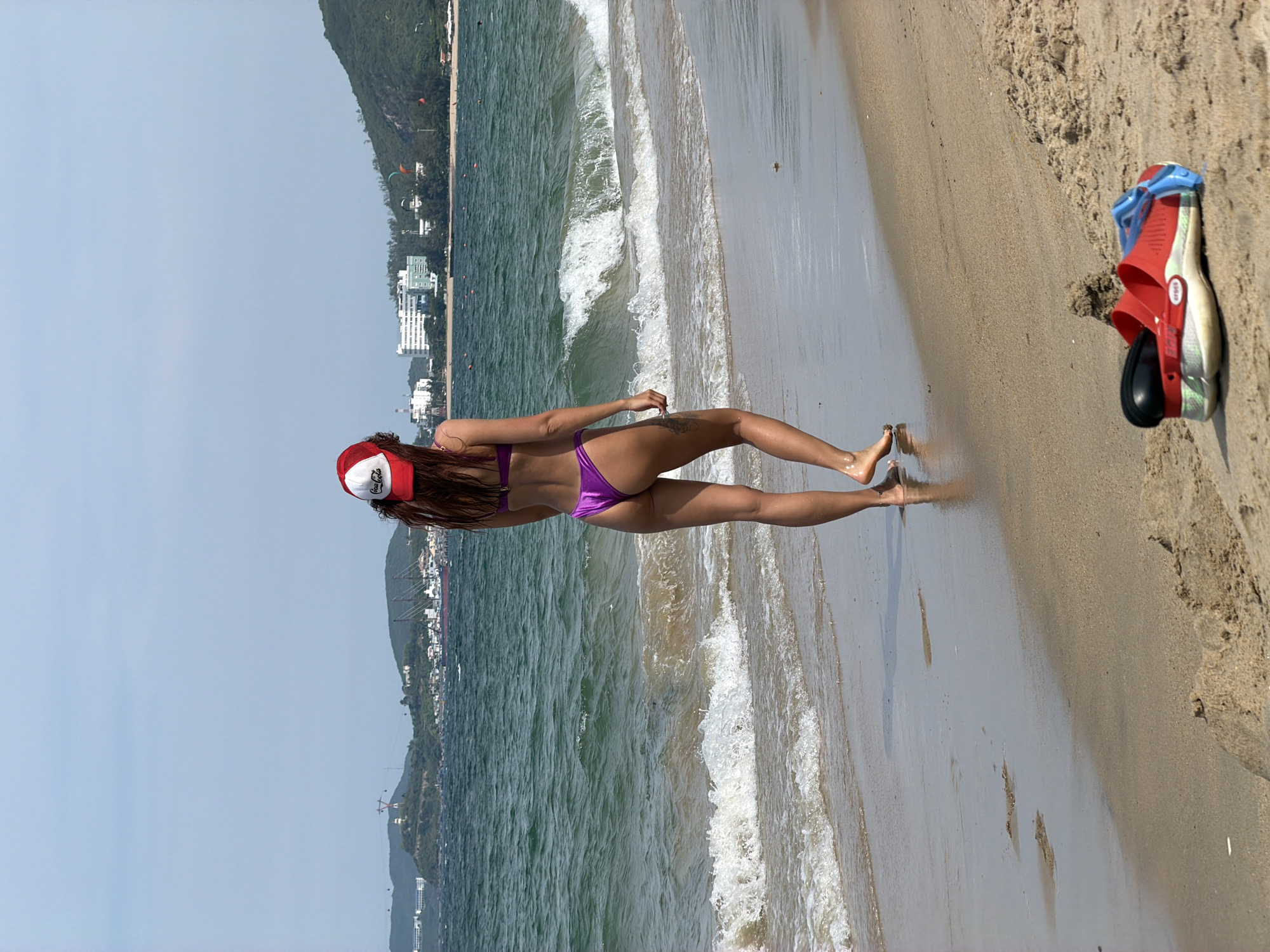A woman wearing a purple bikini and a red and white Coca-Cola hat walks along a sandy beach in Nha Trang Ward, Vietnam. Her back is to the camera as she walks towards the ocean. The water is a greenish-blue with gentle waves lapping at the shore. In the background, a coastline with green hills and some buildings can be seen under a clear blue sky. In the foreground, a pair of red Crocs with blue goggles is placed on the sand.