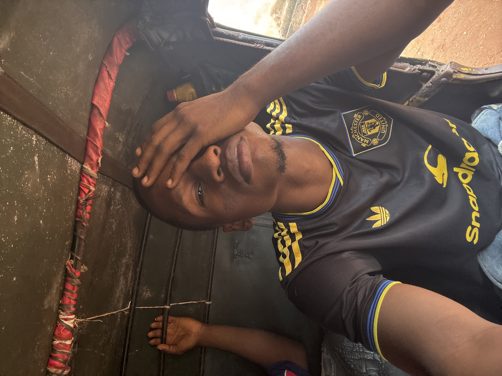 The image shows a man inside a vehicle, likely a "keke" or auto-rickshaw. He is wearing a black Manchester United soccer jersey with yellow Adidas stripes and a "Snapdeal" sponsor logo. He has his hand covering his eyes. The interior of the vehicle appears worn, with a padded red section and metal bars visible. The image was taken in Nkpor, Nigeria.