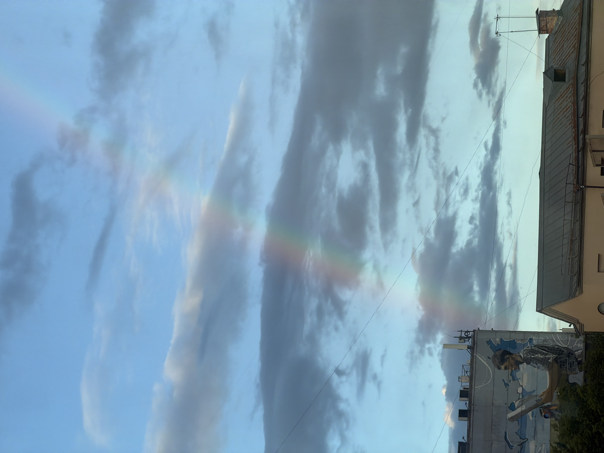 The image shows a partial rainbow in a cloudy sky. The bottom portion of the image contains a portion of a building, and a mural of a man with a map in the background. The image was taken in Moscow, Russia.