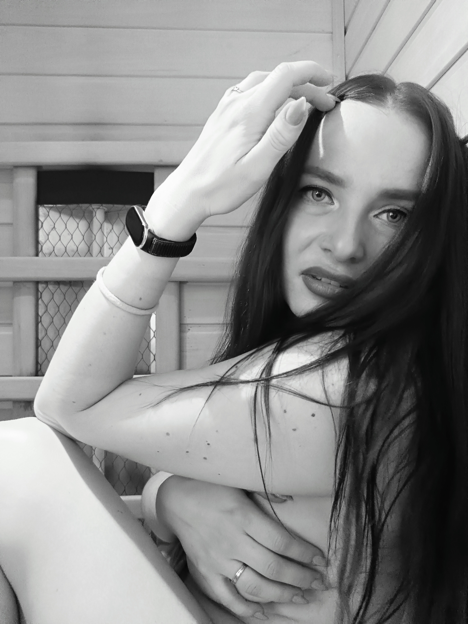 A monochrome medium shot depicts a woman indoors, likely within a structure lined with horizontal wooden planks. She is positioned close to the camera, looking directly at the viewer with a serious expression. Her dark, long hair partially covers her face, and she has dark lipstick. She has fair skin with some visible moles or freckles.
Her left arm is bent, with her hand touching her hair near her forehead. A ring is visible on her finger. On her left wrist, she wears a digital watch and a thin bracelet. Her right arm is across her body, with her hand obscuring her chest.
The background includes a framed, mesh-covered rectangular opening in the wooden structure. There is no discernible information to confirm the location as Chaikovsky, Russia from this image alone.