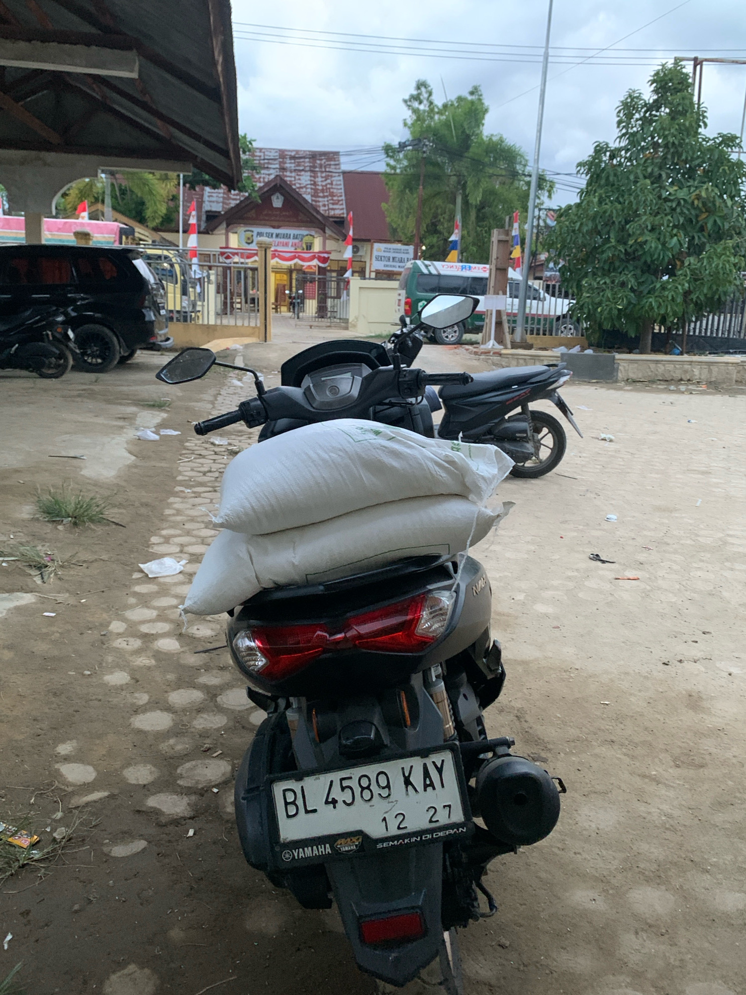 In Pantegurah, Indonesia, a motorcycle is parked on a dirt and paved surface. The motorcycle is black and has two large white sacks stacked on its rear seat. A license plate is visible on the back of the motorcycle, which reads "BL 4589 KAY" and "12 27". A building with red and white flags is in the background, with "POLSEK MUARA BATU" visible on a sign. There are also cars and other motorcycles parked nearby. The overall scene suggests a typical day in a Indonesian town.