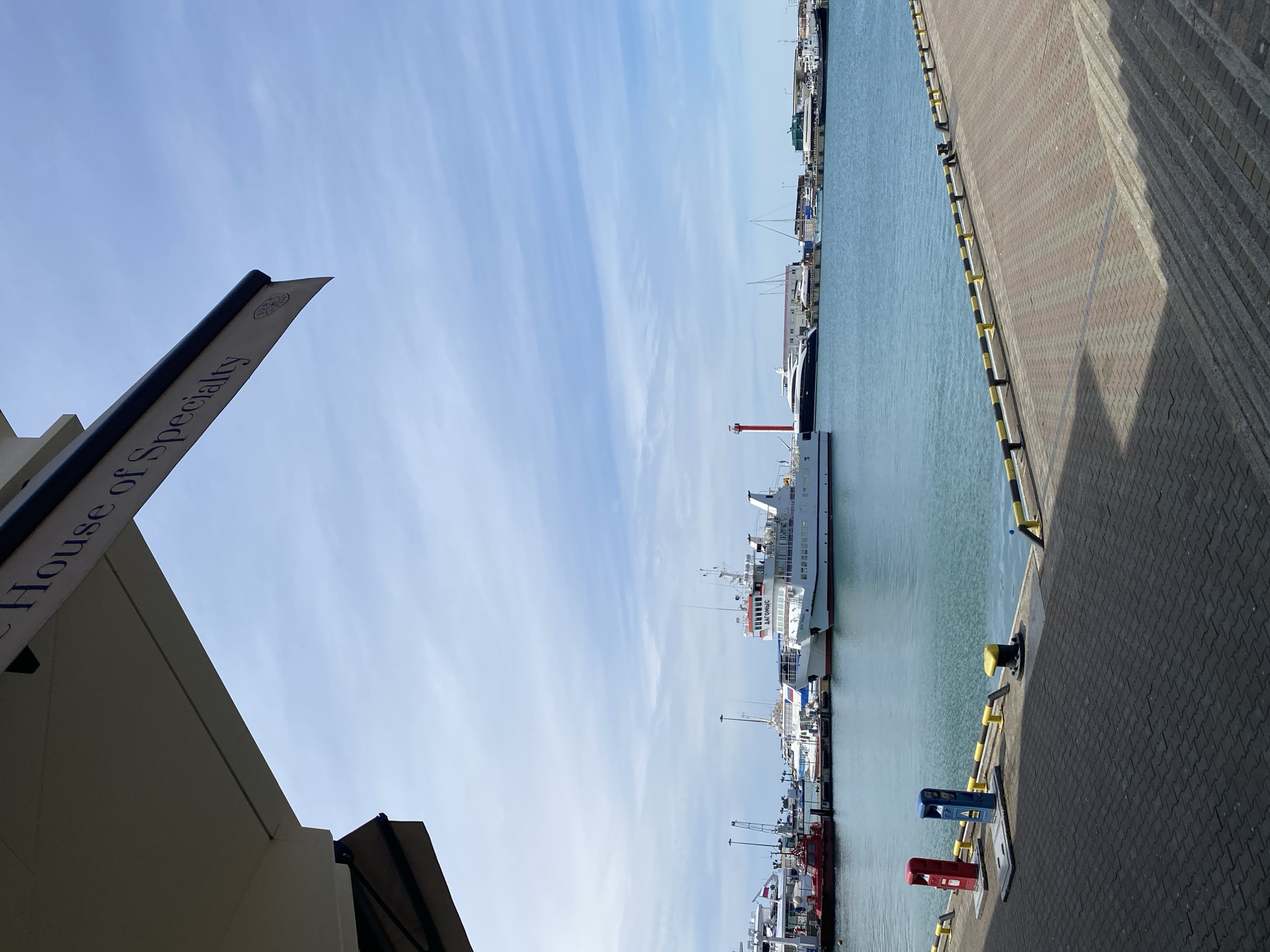 The image shows a waterfront scene in Sochi, Russia. Various boats and ships are docked along a concrete quay with yellow and black safety bumpers. Pavement is made of concrete bricks. A sign is visible in the foreground, reading "House of Specialty." The sky is partly cloudy.