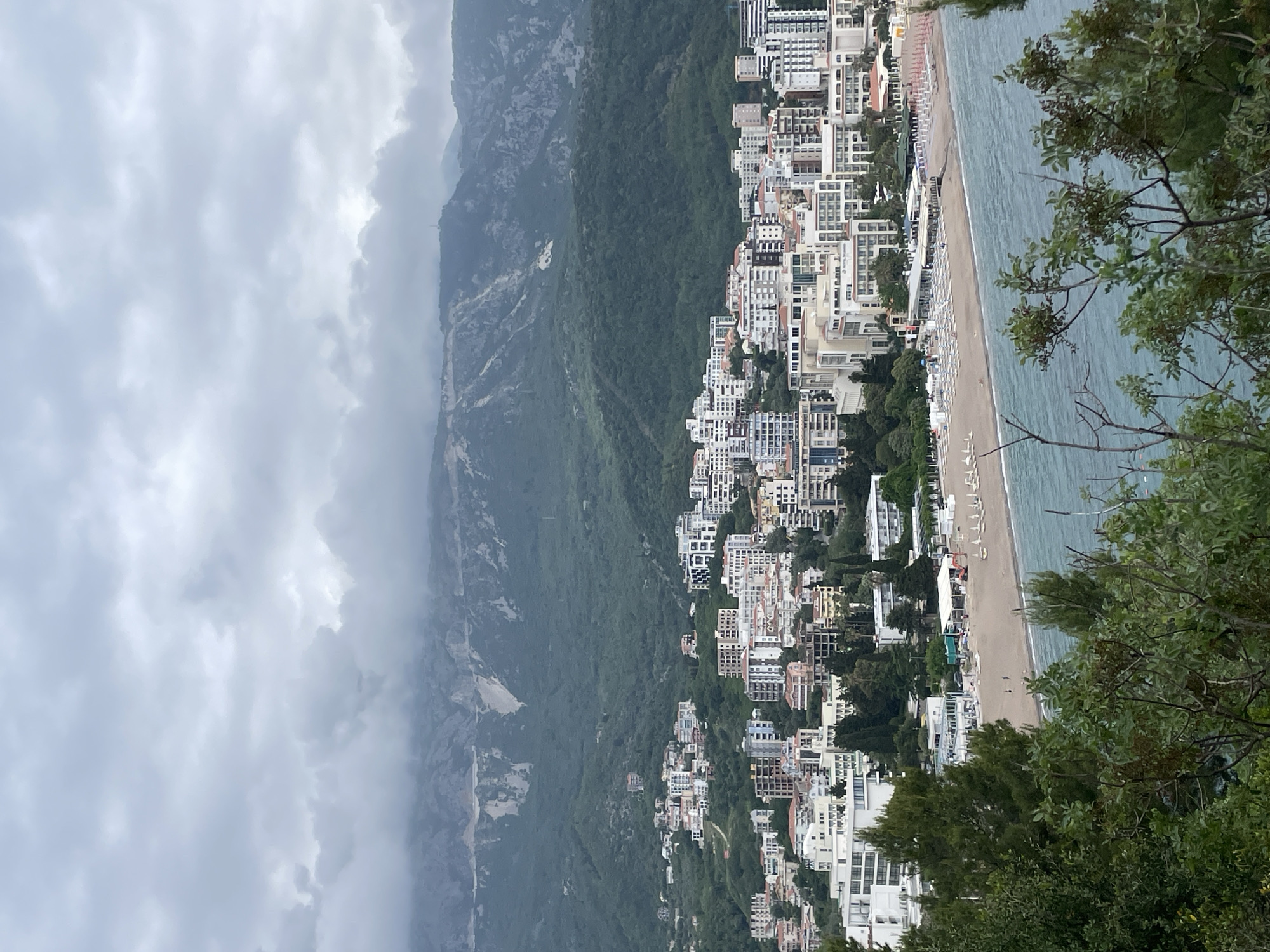The image depicts the coastal town of Rafailovići, Montenegro, on a cloudy day. Tall, modern buildings are clustered along the shoreline, interspersed with greenery. A sandy beach runs along the water's edge, dotted with white beach chairs and a few people. To the left of the town, a large mountain is partially obscured by clouds. The sea is a muted blue-green color. In the foreground, tree branches frame the view from an elevated position.