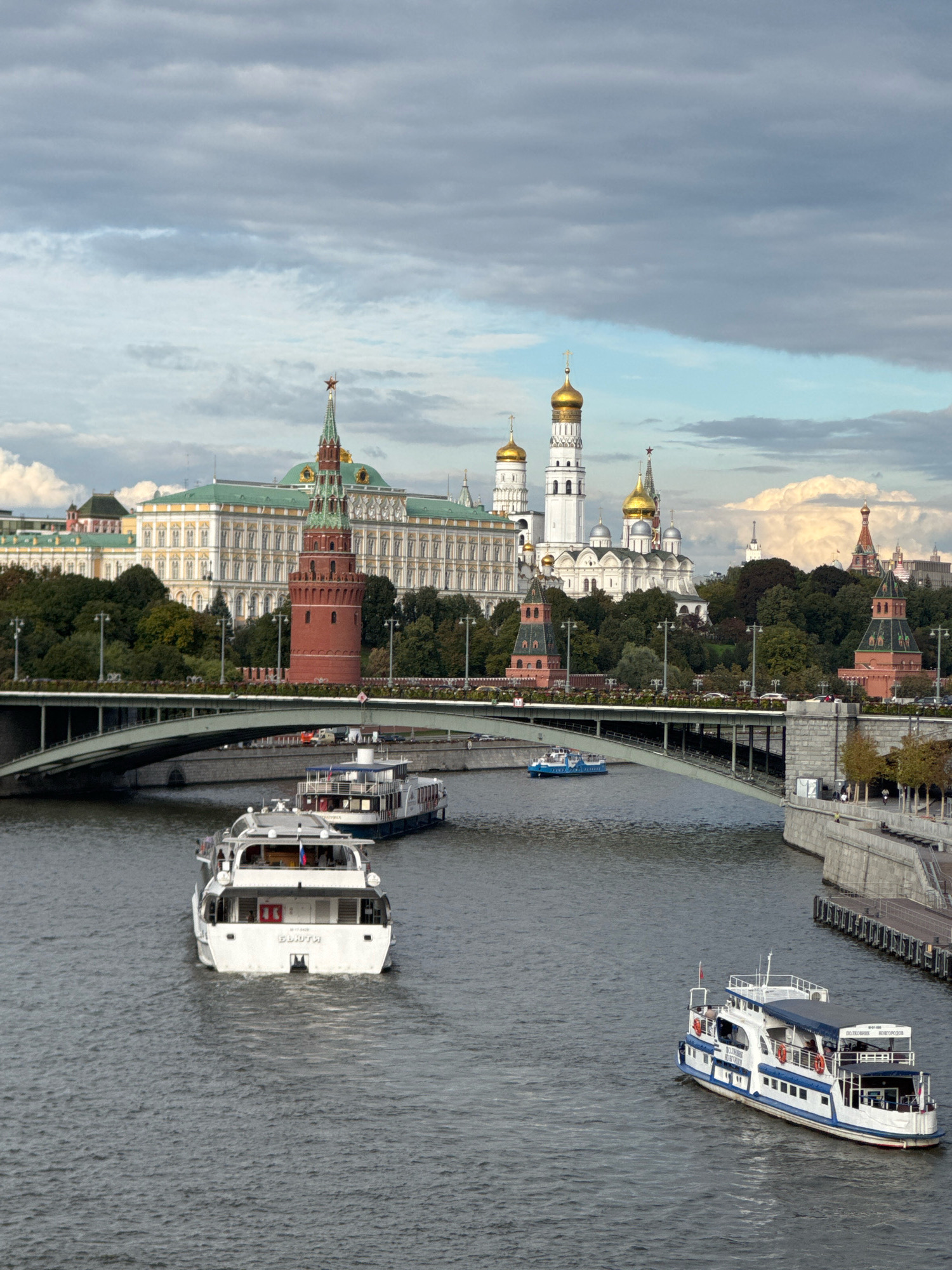 This is a medium-shot, eye-level, vertical, outdoor, daylight photograph taken in Moscow, Russia. The scene captures a wide view of the Moscow River with several tour boats navigating its waters. In the background, the iconic Moscow Kremlin complex is visible, featuring its distinctive red towers and golden domes. A prominent bridge arches over the river, with vehicles and pedestrians visible on its span. The sky is partly cloudy, with a mix of grey and white clouds. The overall atmosphere is serene, with the city's historical landmarks providing a dramatic backdrop to the tranquil river scene.