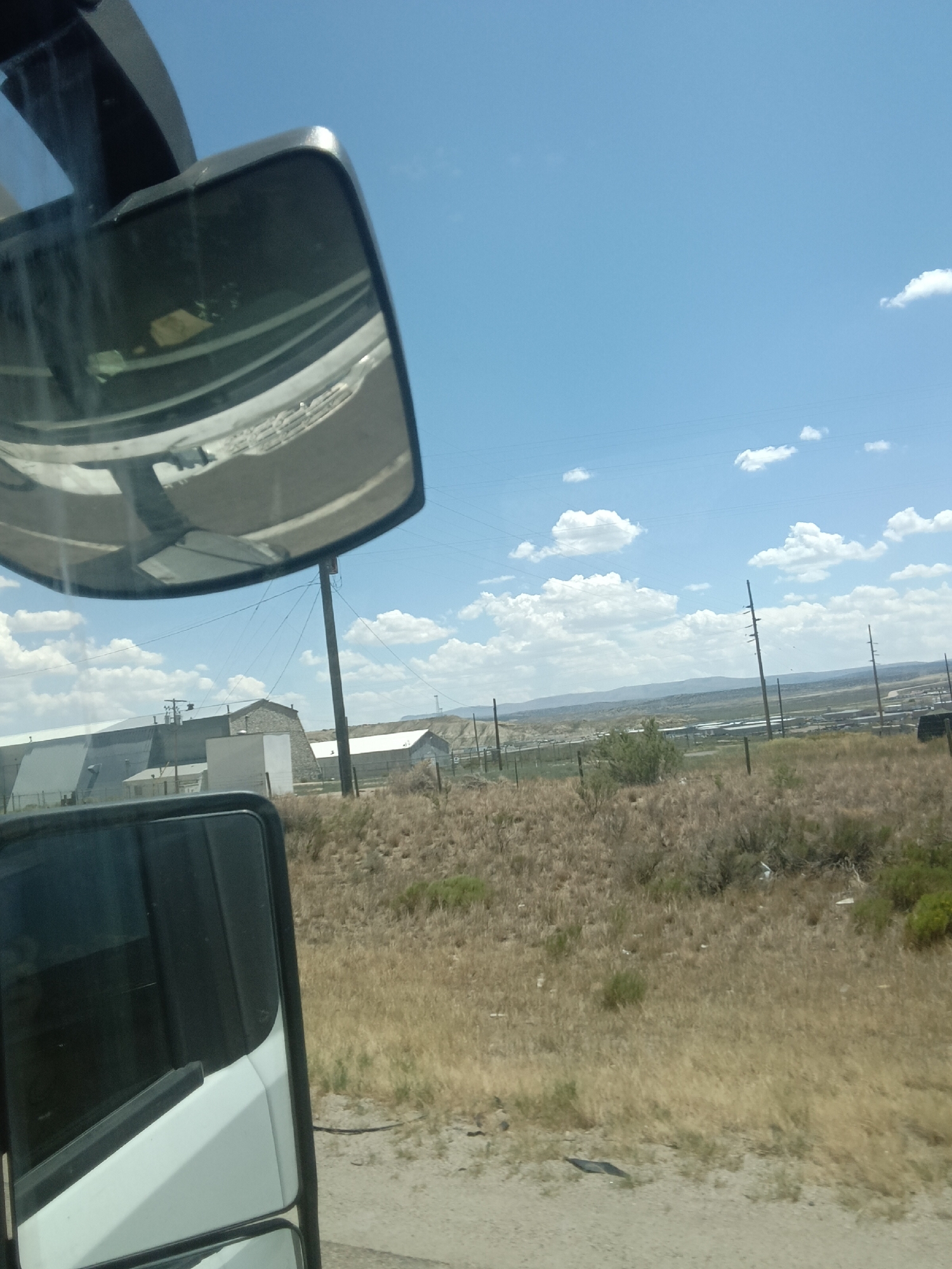 The image depicts a rural landscape with industrial elements under a bright blue sky with scattered white clouds. In the foreground, a side-view mirror of a vehicle reflects a portion of the sky and what appears to be the vehicle's interior, possibly showing the dashboard and windshield. To the left, a white and black vehicle is partially visible.
The middle ground features a collection of industrial buildings, some with large, flat roofs and metallic siding. One building has a stone facade. A tall wooden utility pole stands prominently in the center of the image, with power lines stretching across the scene. Dry, golden-brown grass and sparse green bushes cover the ground, indicative of an arid environment.
In the background, a range of distant hills or mountains is visible, and more utility poles are scattered throughout the landscape. The overall impression is that of an industrial area situated in a semi-arid, open region. The location is identified as Purple Sage, United States of America, which suggests this is a view of the outskirts or an industrial zone within or near that area. No people are visible in the image.