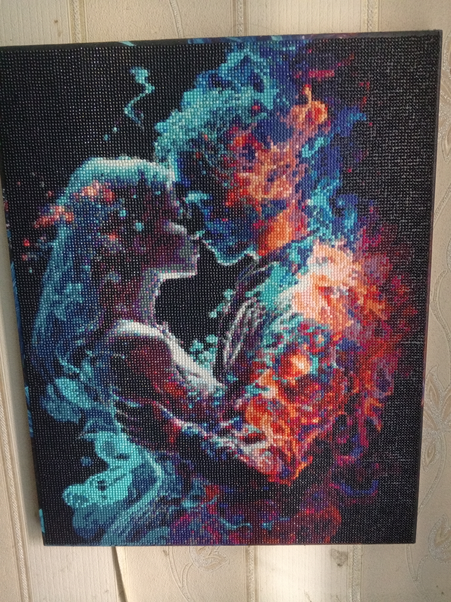 The image shows a diamond painting of two figures in an embrace. The figure on the left appears feminine, depicted in shades of blue and white, while the figure on the right is depicted in shades of red, orange, and blue. The background is black. The location is uncertain, as the question is irrelevant, the image doesn't contain location information, and the location provided is not relevant for the question.