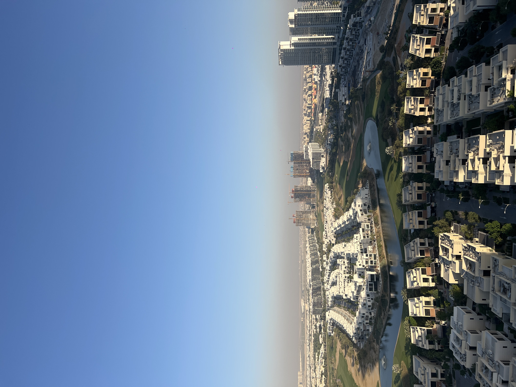 The aerial photograph shows an urban landscape in Dubai, United Arab Emirates. The sky is clear blue. The urban area includes white villas arranged along the right side of the frame near a canal or waterway. Further away there are green golf courses and larger buildings. Some buildings are under construction. The picture has been rotated 90 degrees counterclockwise.
