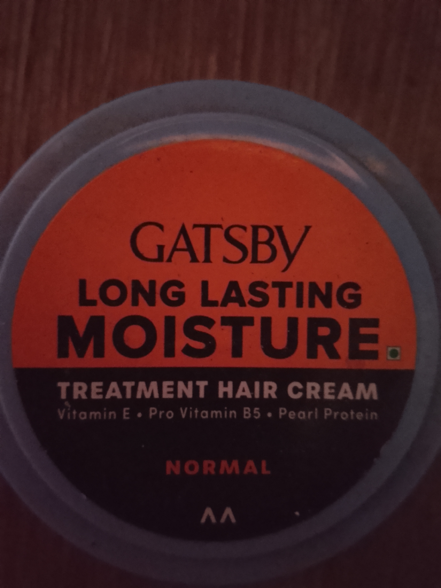The image shows a top-down view of a Gatsby Long Lasting Moisture Treatment Hair Cream jar. The lid is orange with black text. The text reads "GATSBY" in large script at the top, followed by "LONG LASTING MOISTURE" in all caps. Below this, it says "TREATMENT HAIR CREAM" with "Vitamin E • Pro Vitamin B5 • Pearl Protein" in smaller text underneath. At the bottom of the orange label, the word "NORMAL" is written in all caps. The jar itself appears to be a light blue or gray color and is resting on a dark brown, wood-grain surface, possibly a table or floor. There are no people or animals visible in the image, nor any specific landmarks that would indicate the city of Kasiman, Indonesia.