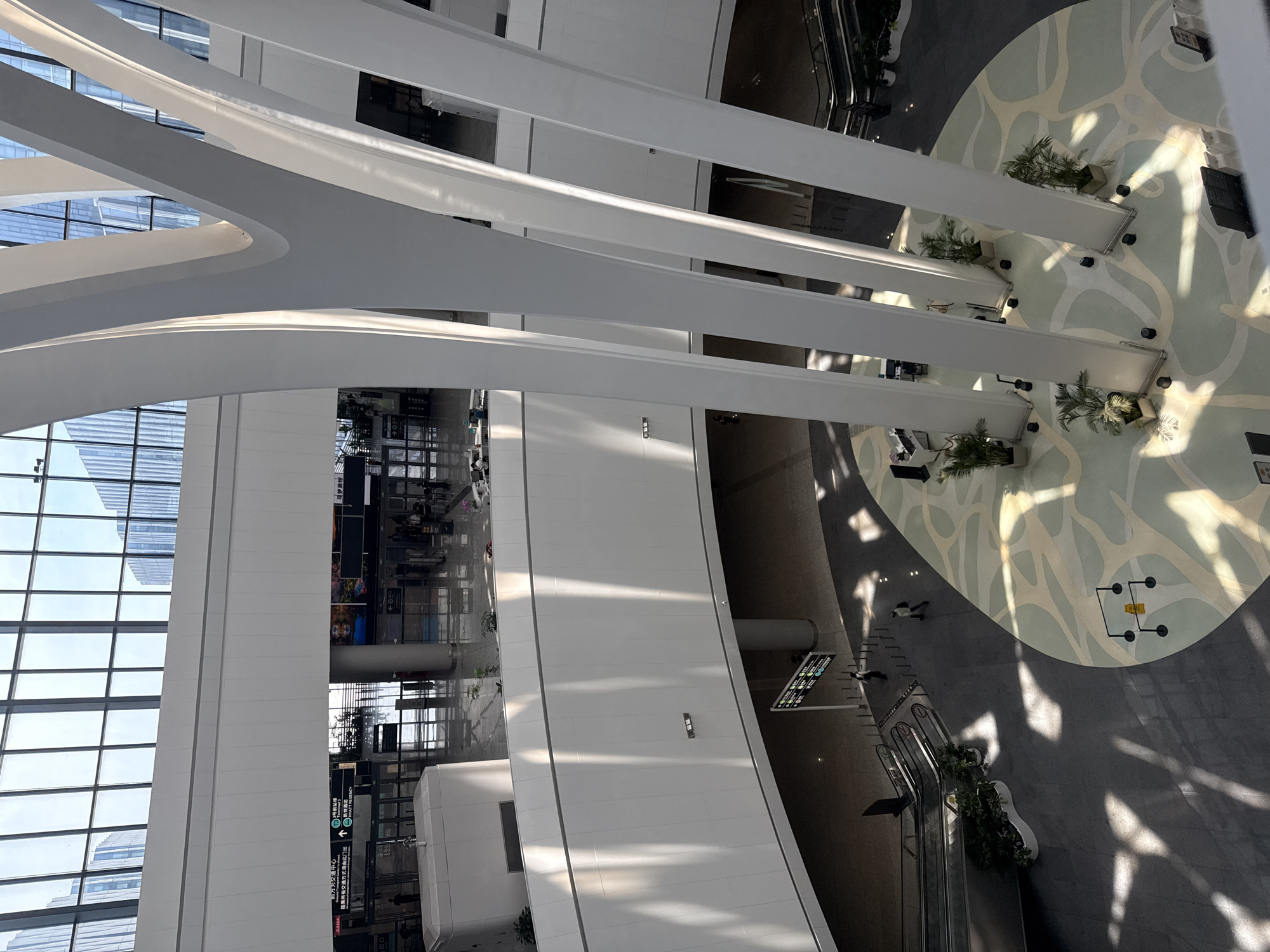 The image shows the interior of a modern, multi-level building, likely an airport or transportation hub, in Xiaoshan District, China. The space is characterized by its vastness, natural light filtering through a large glass ceiling, and distinctive architectural features. Tall, white, curved columns dominate the central area, converging towards the ceiling.
On the lower level, a circular floor design with organic, flowing patterns in muted colors creates a visually engaging space. Several potted plants are strategically placed around the base of the columns. Escalators are visible on the left side of the frame, leading to different levels, and a few individuals are walking on the floor. Signage is present, including directional signs and what appears to be information kiosks.
The middle level features a curved white wall with a series of horizontal windows or panels, and more light streams in from the upper levels. Signs indicating "Terminal 3" and "HYATT REGENCY" are visible on this level, suggesting a connection to travel and accommodation. Further back, through large glass doors, a concourse area with more people and what appear to be check-in counters or gates can be seen.
The upper level, visible through the expansive glass ceiling, shows reflections of tall buildings, indicating the structure is located in an urban environment. The overall impression is one of spaciousness, modernity, and efficiency.