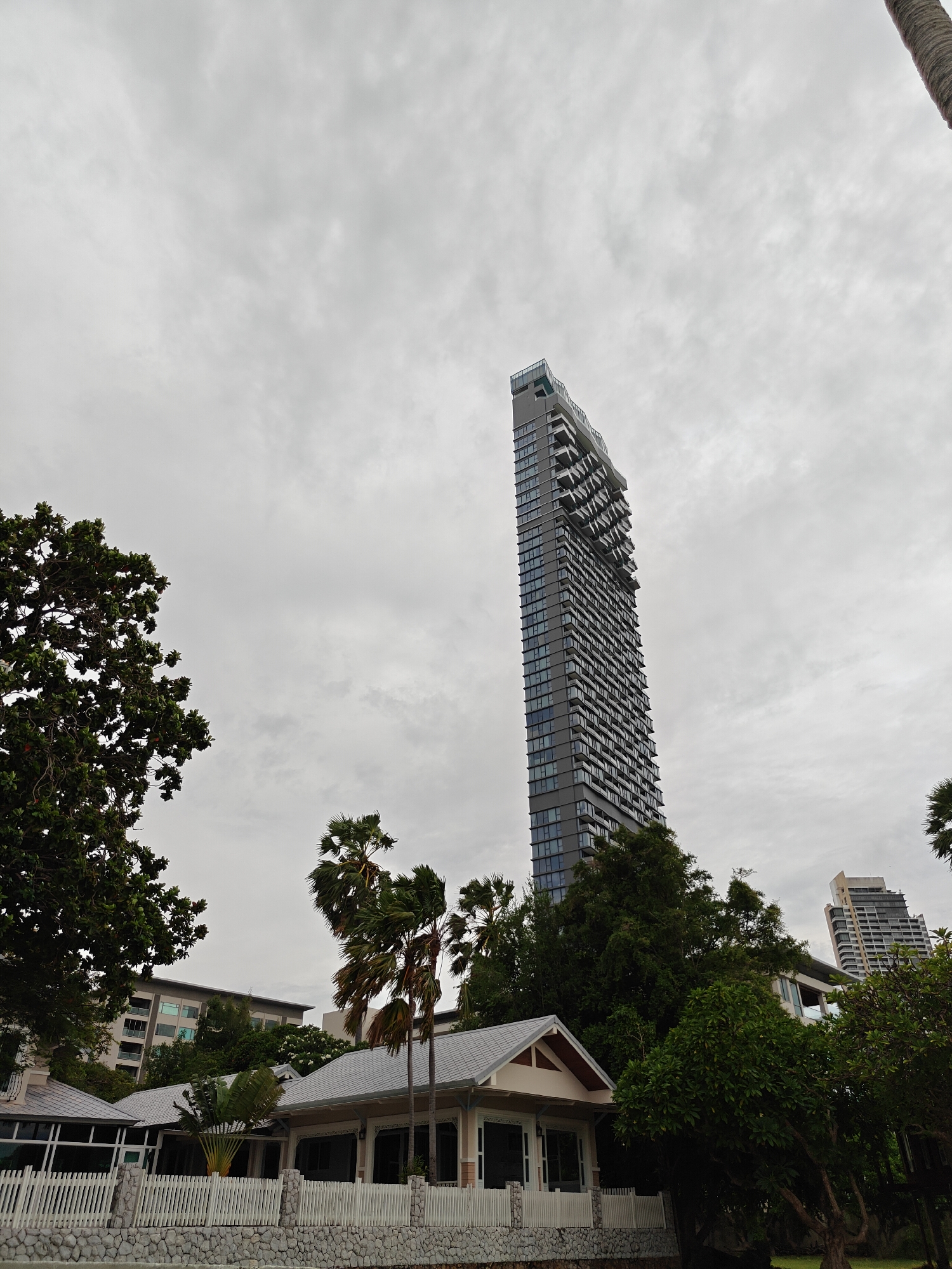 The image shows a cluster of buildings in Pattaya City, Thailand, under a cloudy sky. A prominent, tall, modern apartment building with a unique, angled top and numerous balconies dominates the skyline. In the foreground, a low-rise building with a pitched roof, white picket fence, and stone base suggests a resort or residential area. Palm trees and lush green foliage are scattered throughout the scene, indicating a tropical climate. In the background, another, shorter apartment building is visible. The overall impression is one of a coastal urban environment with a mix of modern and traditional architecture, set against a muted, overcast sky.