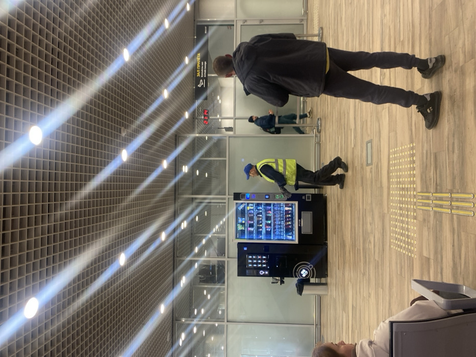 The scene depicts an interior view of an Arrivals Lounge ("ЗАЛ ПРИЛЁТА") at 21:14, located in Новая Казмаска, Russia. The environment features a high ceiling with a gray grid pattern, illuminated by numerous recessed circular lights that cast distinct diagonal light rays. The floor is covered with light-colored wood-look laminate, incorporating yellow tactile paving strips. Glass partitions form the background.
Two dark-colored vending machines are positioned centrally against a light wall. The left machine appears to be a beverage dispenser, while the right machine, internally lit with blue light, displays various packaged snacks and drinks. A stainless steel trash receptacle stands to the left of these machines.
Four individuals are present. In the left foreground, the head and shoulder of a seated person with light hair are visible, facing right. In the mid-ground, a male wearing a blue cap, a bright yellow high-visibility vest, and dark trousers is standing in front of the snack vending machine, with his hands near its front, suggesting interaction for servicing or stocking. To his right, a second male stands with his back to the camera, dressed in a dark hooded jacket, dark trousers, and dark athletic shoes, with his left hand slightly raised. Further in the background, a third male wearing a dark jacket and a backpack is walking from left to right past the "Arrivals Lounge" sign.
