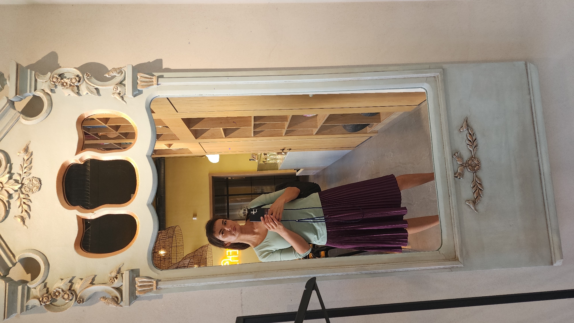 A woman is reflected in a decorative mirror. The mirror has an ornate design with three oval cutouts and floral ornamentation. The reflection shows a room with wooden shelving, yellow walls, and a neon sign. The woman is wearing a light green shirt and a dark purple pleated skirt. She is holding a smartphone in front of her face. A black chair is visible in the foreground of the reflection. The photograph appears to have been taken indoors. There is no information indicating it is taken in Smolensk, Russia.