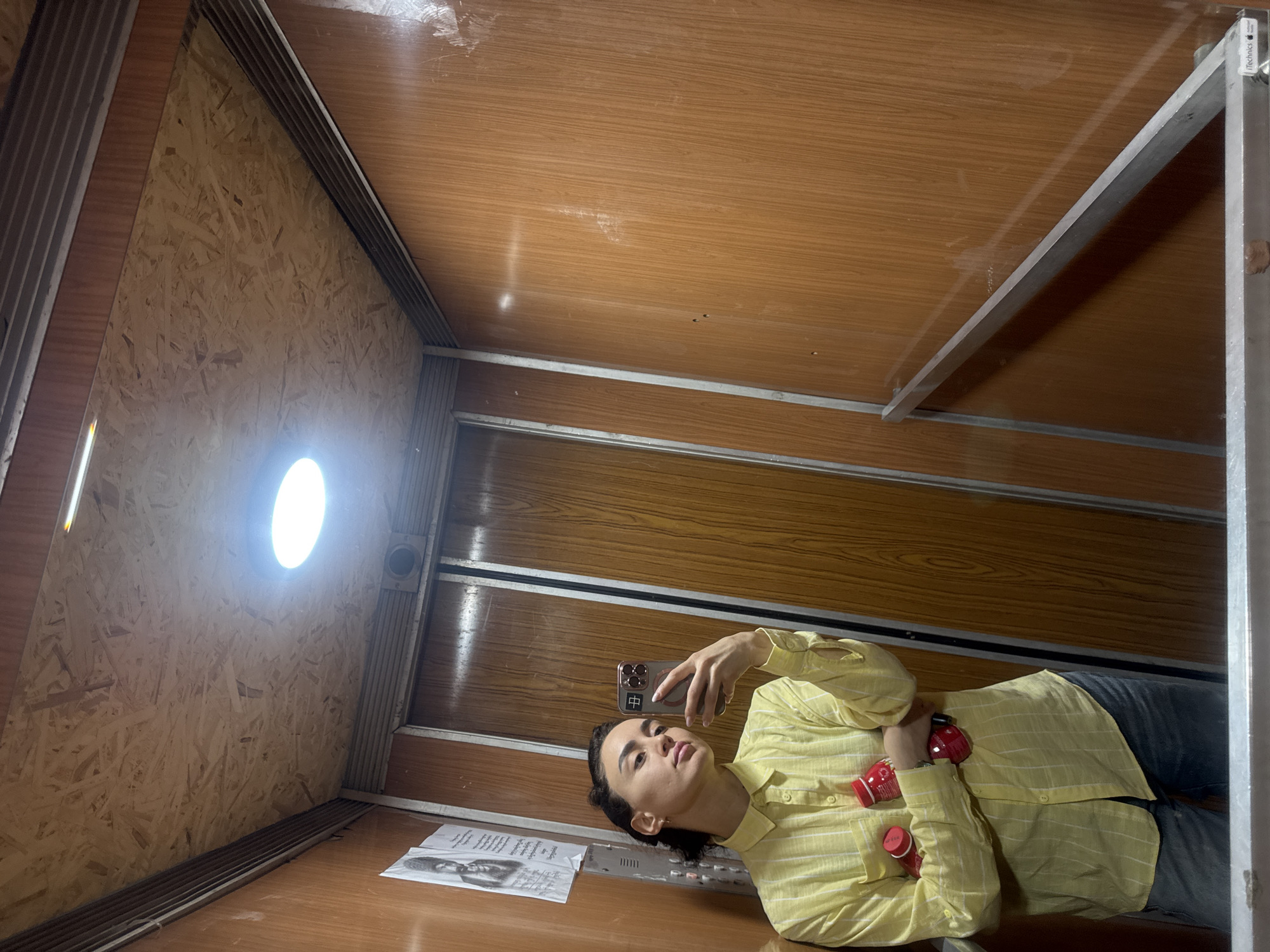 A person is lying on the floor of what appears to be an elevator or small room, taking a selfie with a smartphone. The person is wearing a light yellow striped shirt and dark jeans. They are holding the phone in their right hand. There is a red object, possibly a container, near their chest.
On the floor next to the person is a piece of paper with an image and text. The walls of the enclosure are made of plywood on one side and a wood-grained paneling on the other. There's a circular light fixture on the plywood wall. The overall orientation suggests the camera is angled upward. The location is in Tbilisi, Georgia.