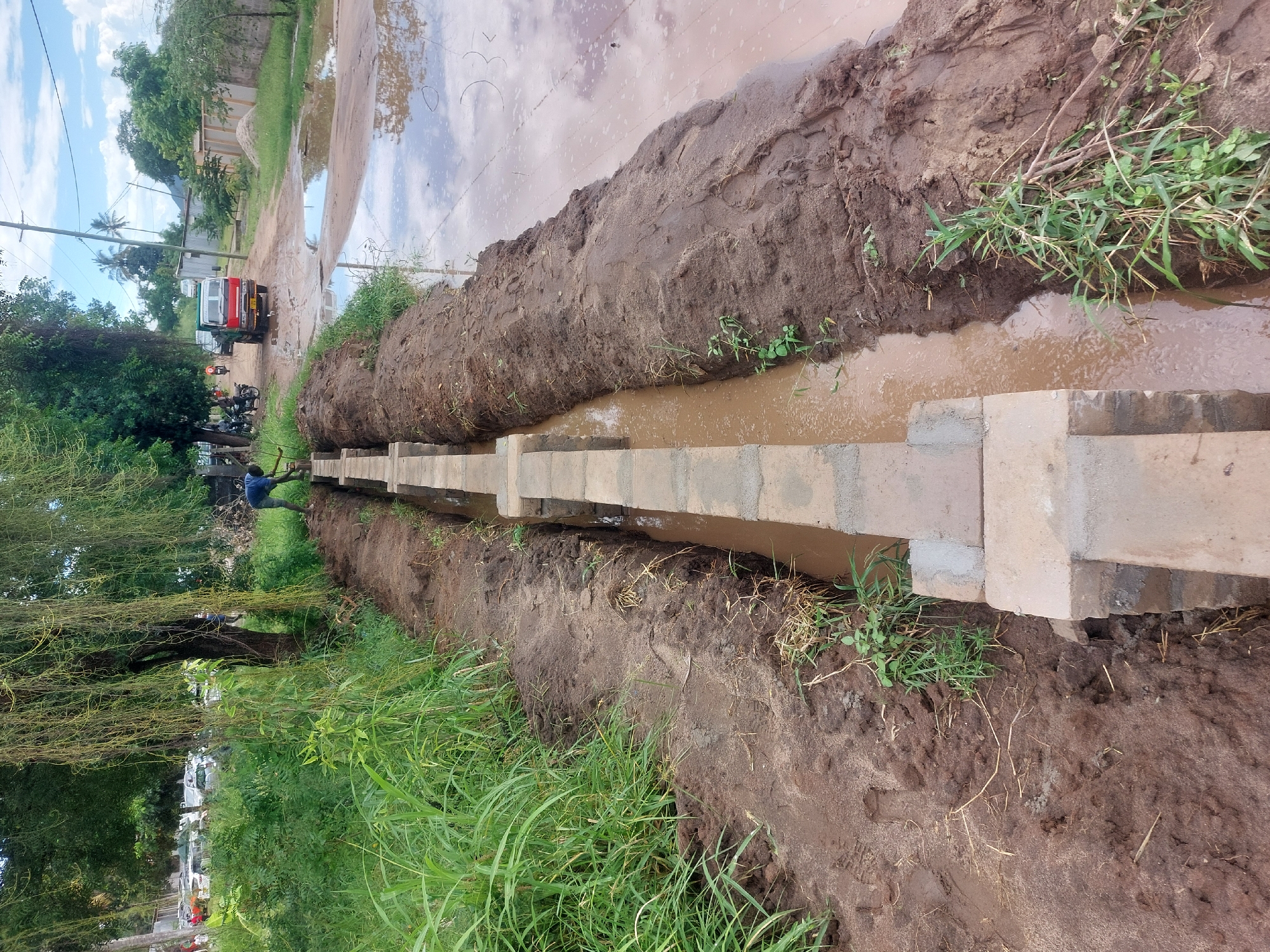 The image depicts a newly constructed drainage canal in a city environment, likely in Dar es-Salaam, Tanzania. The canal is constructed from gray concrete blocks, forming a straight line through a muddy ditch. Water is present inside the canal and along the edges. The surrounding area consists of dirt piles and grassy vegetation. In the background, a flooded street reflects the sky. A person is visible working on the canal construction towards the left side of the image, near a tree. Other vehicles and buildings can be seen in the distance. The overall scene suggests an ongoing construction or infrastructure development project in a potentially flood-prone area.