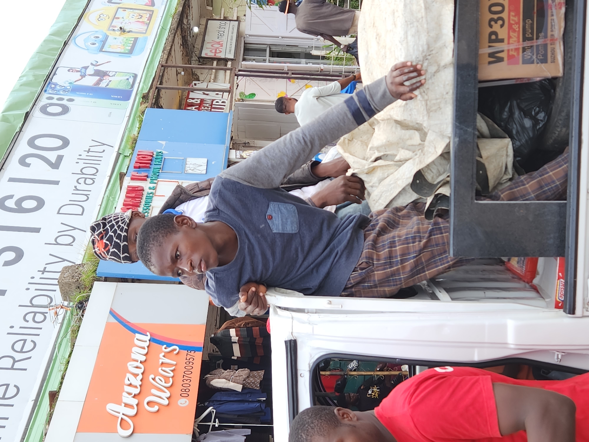 The image depicts a street scene in Jos, Nigeria. In the foreground, a young man wearing a grey long-sleeved shirt and brown plaid pants is leaning out of the back of a white pick-up truck, holding onto the edge of the truck bed. He is looking to his left. Part of another person wearing a red t-shirt is visible on the left side of the frame. The truck bed is filled with various items, including a large piece of beige tarpaulin and black trash bags. A cardboard box labeled "WP301 FM&T Water pump" is visible in the lower right corner of the truck bed.
Behind the truck, there are several shops. A prominent sign reads "Arizona Wears" with a phone number "08037009575". Another sign, partially visible, says "Oman Action Accessories & Phone Parts". Further in the background, another sign indicates "Felak meat shop". People are seen walking in front of the shops, including a man in a light green shirt and another in a brown t-shirt. Overhead, a large banner with text like "Reliability by Durability" and images of mobile phones is displayed. The overall environment suggests a busy commercial area.