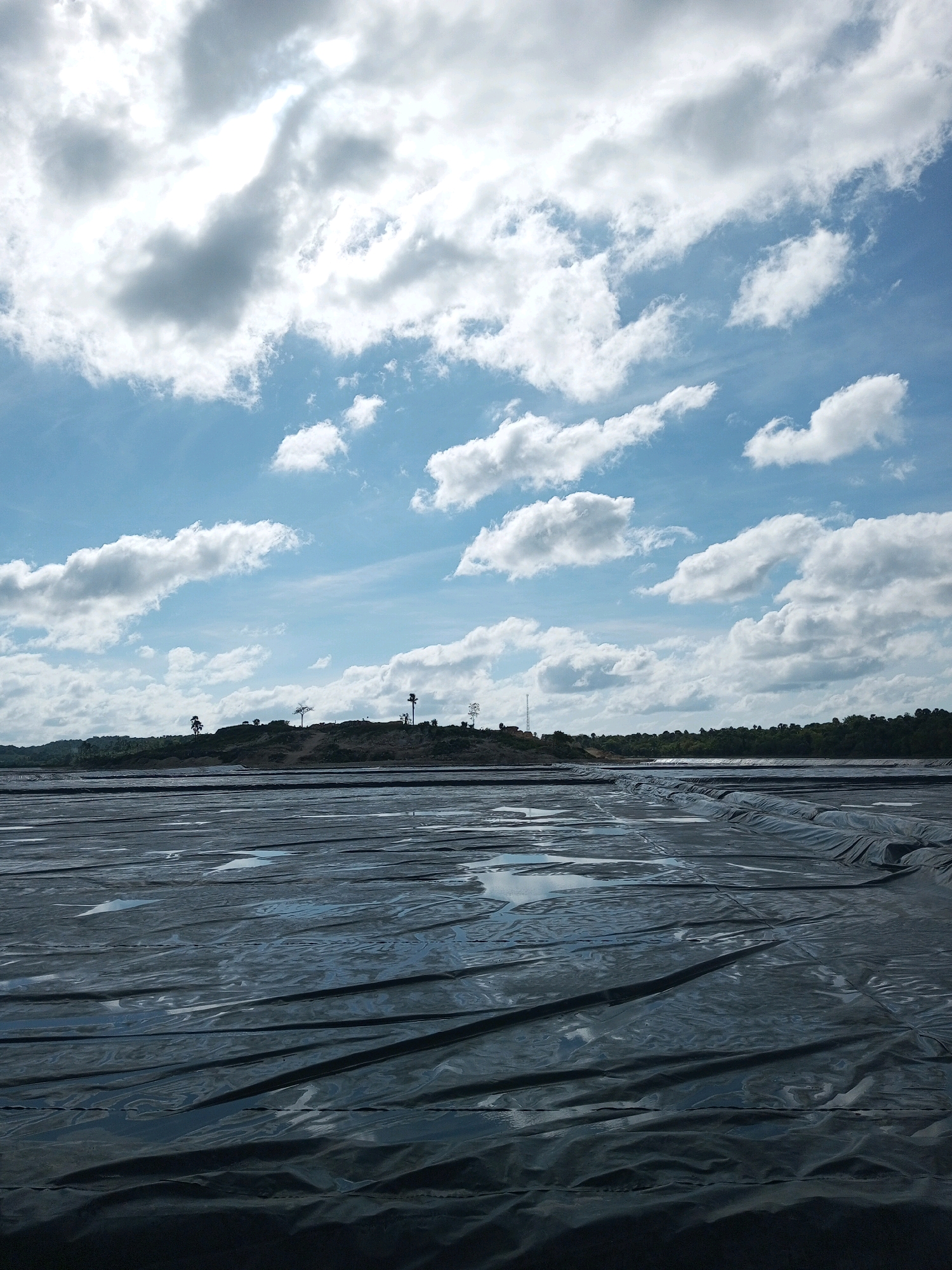An outdoor, wide-angle shot captures a landscape under a bright, cloudy sky with a distant horizon of trees and hills. The foreground is dominated by a large, dark, tarp-like material spread across the ground, with visible folds and ripples. Puddles of water are scattered across the surface of this material. In the middle ground, a gentle, sloping hill is visible, dotted with sparse trees and what appear to be man-made structures or mounds. The sky above is a vibrant blue, filled with numerous white, fluffy clouds, some of which are illuminated by the sun, creating areas of bright light and softer shadows. The overall scene suggests an outdoor construction site or a large-scale containment area. The location of Matasio, Indonesia is not visually discernible from the image itself.