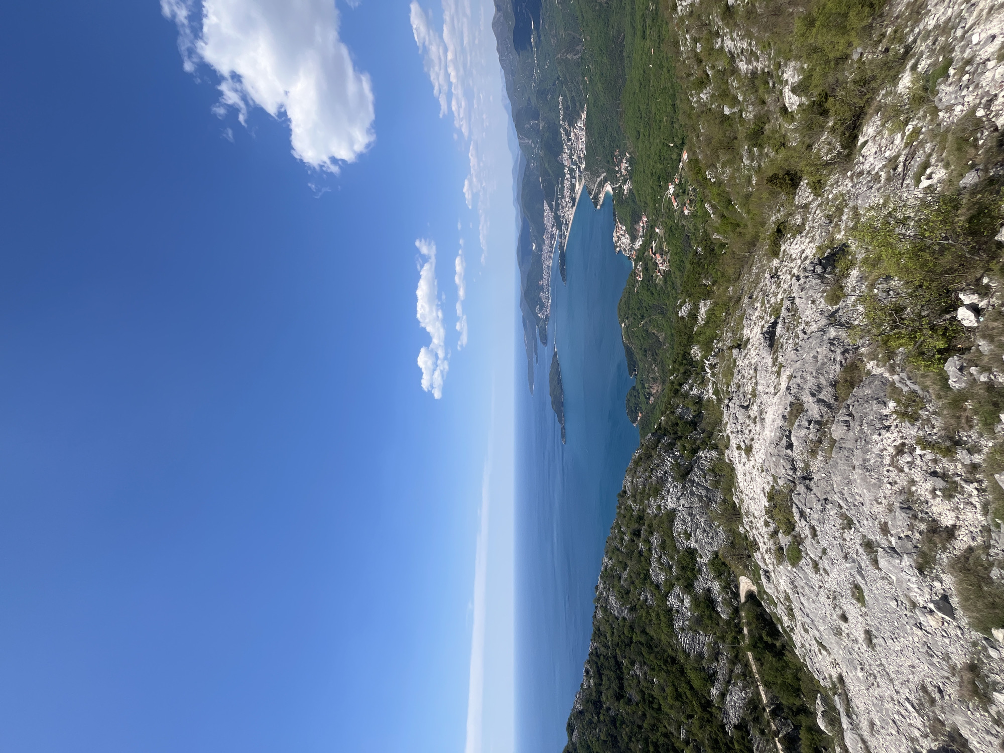 The image captures a high-angle view along the coast of Vrijesno, Montenegro. The azure waters of the Adriatic Sea meet the rocky coastline, and a stretch of the town is visible along the shore. A mountain slope, covered with sparse vegetation and exposed rock, dominates the right side of the frame. The sky is a clear blue with scattered, fluffy white clouds on the left.