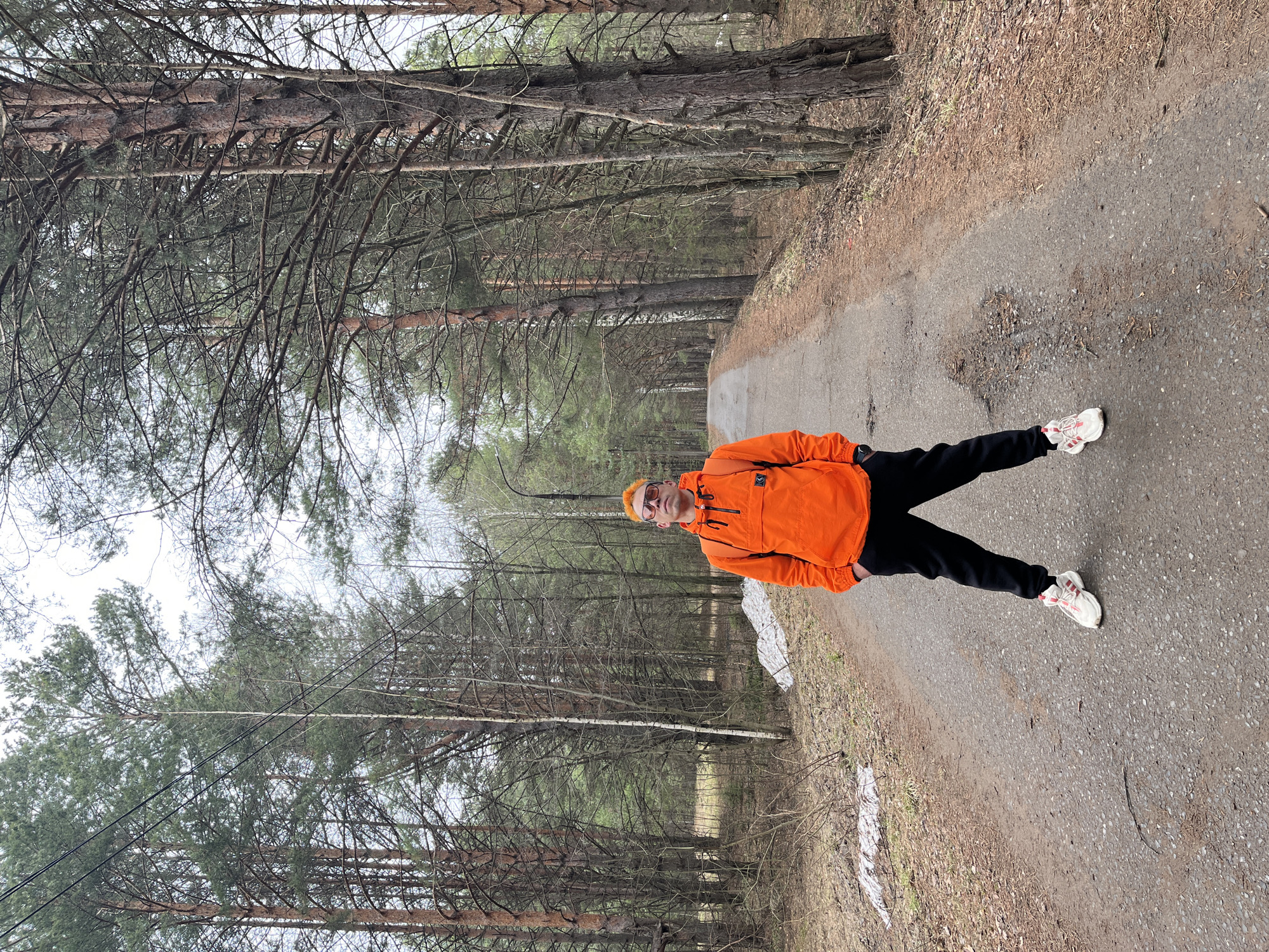 The image shows a person standing on a paved path in a wooded area, possibly in the city of Chaikovsky, Russia. The individual is wearing an orange jacket, black pants, and white shoes with red accents. They have an orange hat and sunglasses. The path is surrounded by tall trees. Snow is visible alongside the path. The person is standing with their legs spread wide apart.