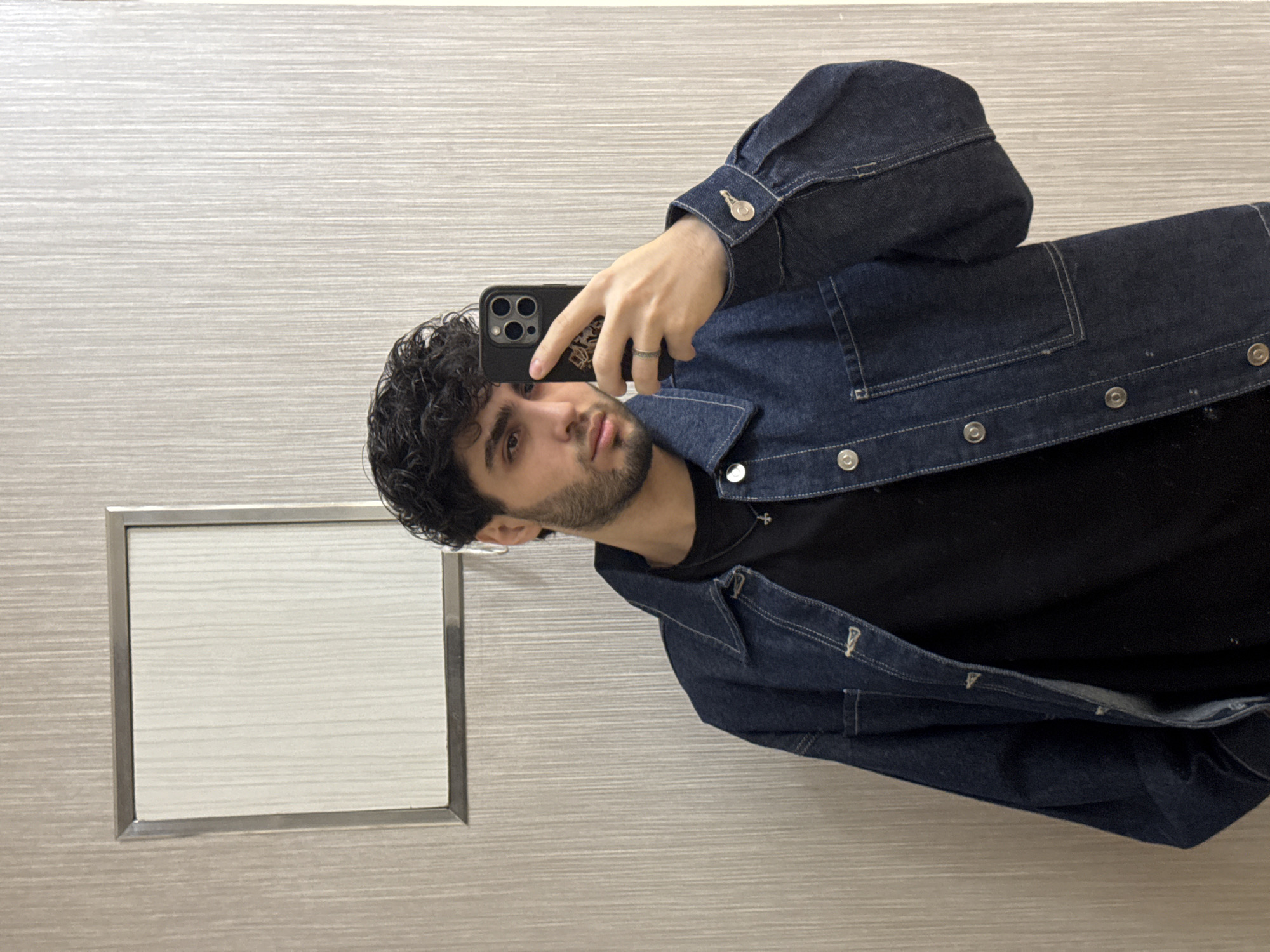 A young man is taking a selfie in what appears to be a bathroom. He has dark curly hair, a beard, and is wearing a dark denim jacket over a black t-shirt. He is holding a black smartphone with a ring on his finger. The background shows a light-colored wall with horizontal lines and a silver-framed rectangular panel, possibly a mirror. The image does not provide enough information to determine the location in Yerevan, Armenia.