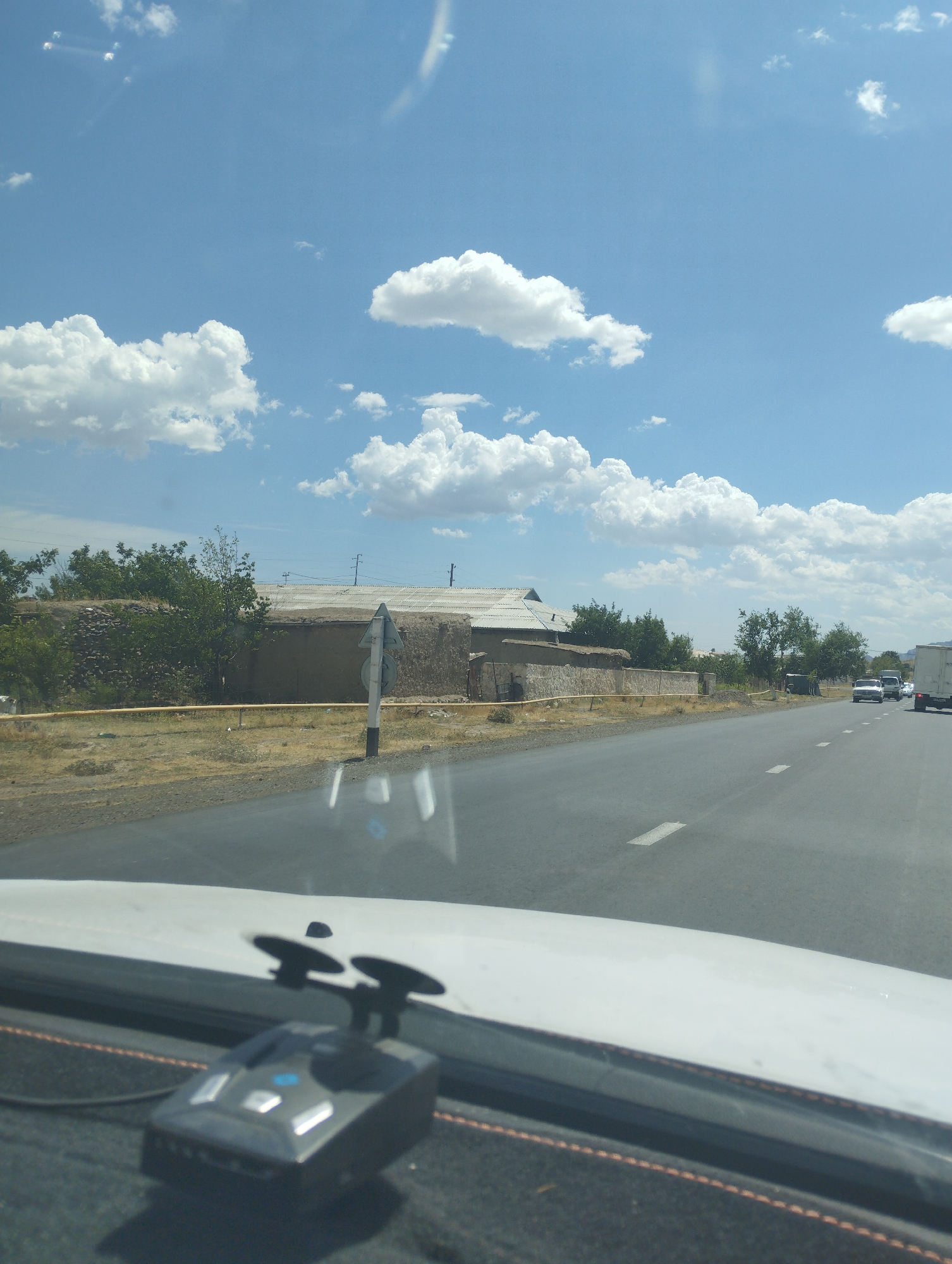 The image depicts a view from inside a vehicle, looking out through the windshield at a road in Asmansay, Uzbekistan. The sky is a clear blue with scattered white cumulus clouds. On the right side of the road, there are several vehicles, including what appears to be a white truck and other cars. The road itself is asphalt with white lane markings. To the left of the road, there is a dirt or gravel embankment with sparse vegetation. Further back, there are low-lying buildings with flat roofs, constructed of mud or adobe. Some trees and bushes are visible in the background, and there are power lines and poles. In the foreground, on the dashboard of the vehicle, a black radar detector with two suction cups is visible. The overall scene suggests a sunny day in a rural or semi-rural area.