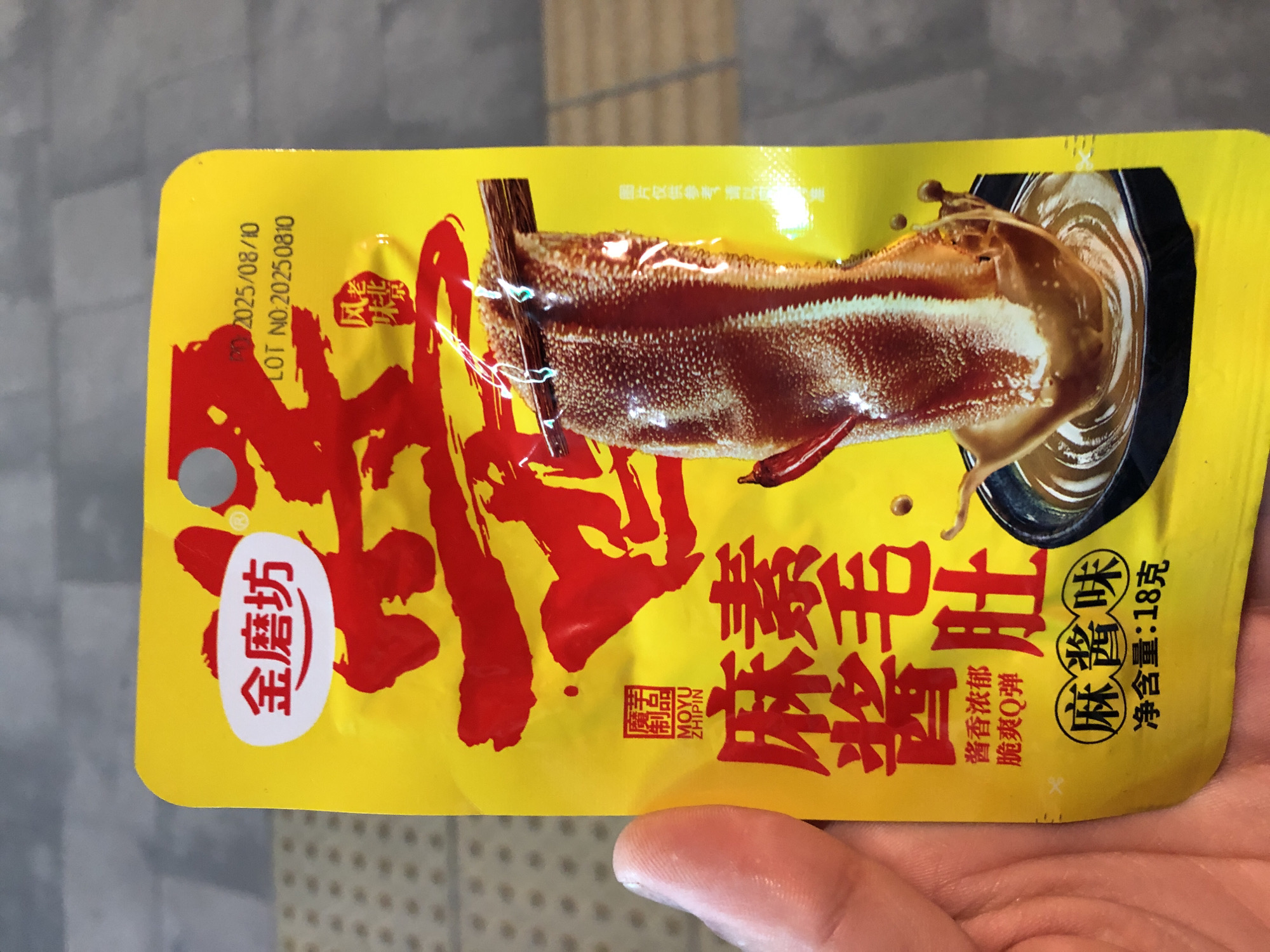 An overhead shot captures a hand holding a yellow food package with bold red Chinese characters and an image of a piece of tripe drenched in sauce. The package also displays text in English. The background is a blurry, tiled surface.