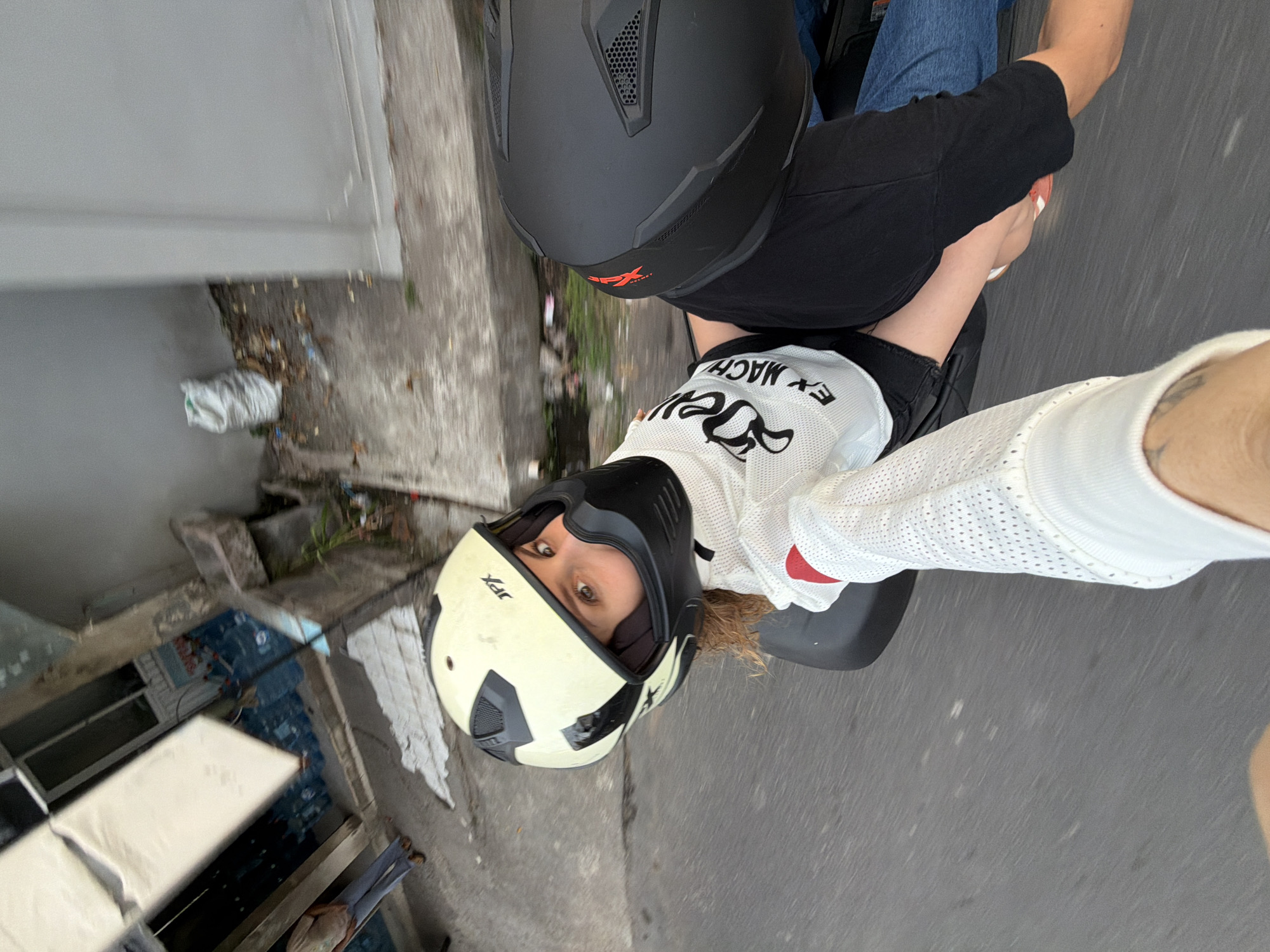 A first-person perspective shot shows two individuals on a motorcycle. The foreground is dominated by the arm and hand of the person taking the photo, wearing a white mesh glove with red accents. The individual in the foreground, likely the rider, is wearing a light cream-colored open-face helmet with black accents and a black chin guard, with visible blonde curly hair. They are wearing a white mesh top with black lettering that reads "EX MACHINE". The second individual, the passenger, is partially visible, wearing a black helmet and sitting behind the rider. They are wearing a black shirt and blue jeans. The motorcycle's seat and frame are visible.
The background depicts a street scene in what appears to be a developing urban environment. To the left, there is a building with large white panels and visible blue water bottles stacked inside. A person in a light-colored shirt and blue pants stands near the entrance. The ground is paved, and a concrete drainage ditch runs alongside the street, filled with debris, leaves, and a white plastic bag. The scene is captured in motion, indicated by the blurred asphalt of the road. The location is identified as North Kuta, Indonesia.