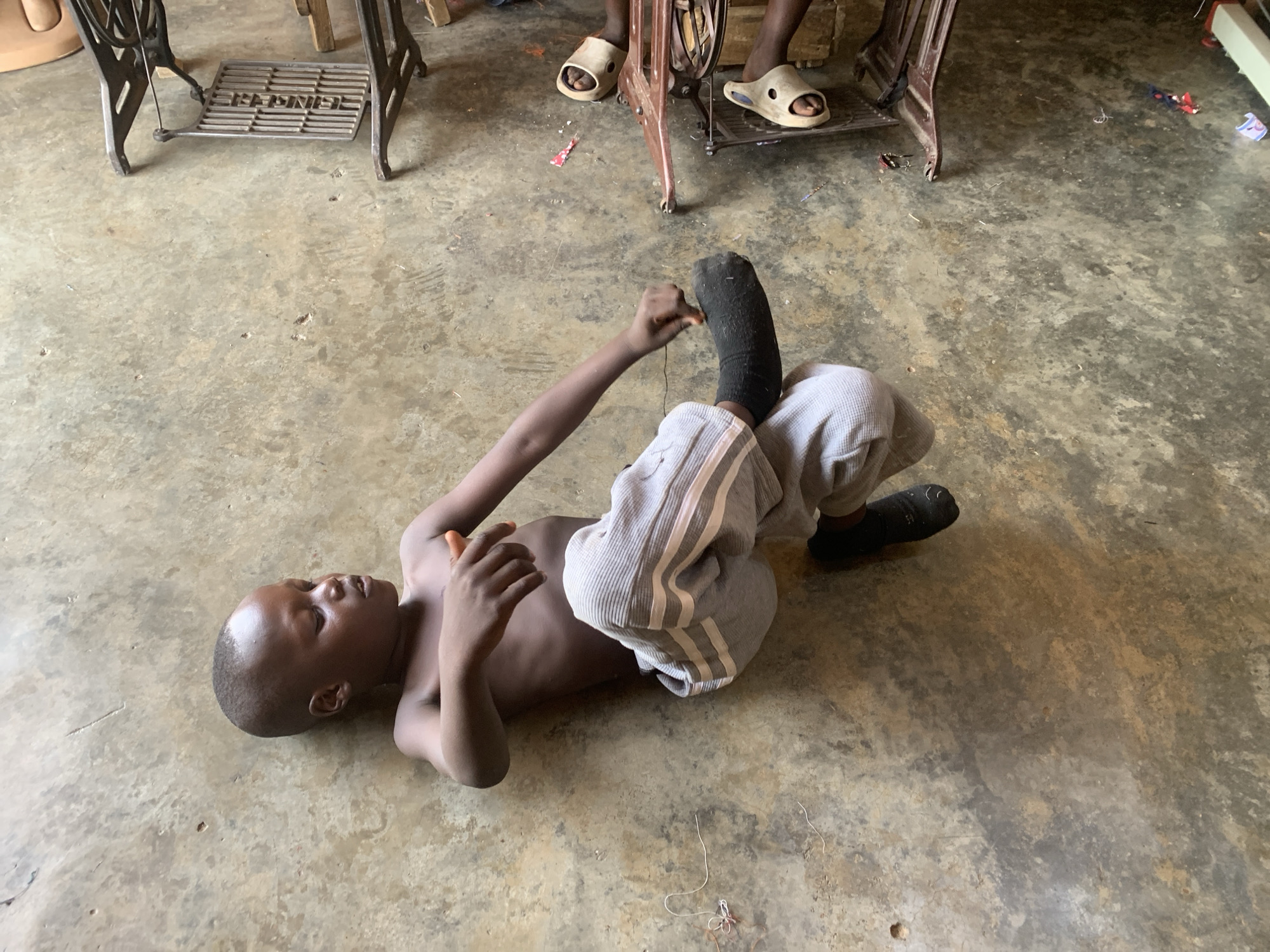 A shirtless, dark-skinned boy with a right leg prosthesis lies on a concrete floor indoors. He wears grey sweatpants with white stripes, a black sock on his prosthetic leg, and one black shoe on his other foot. The boy is manipulating the prosthesis with his hands. Two sewing machines are partially visible in the background. Two adults sit at the sewing machines, wearing sandals. The precise location within the city of Jos, Nigeria is indiscernible.