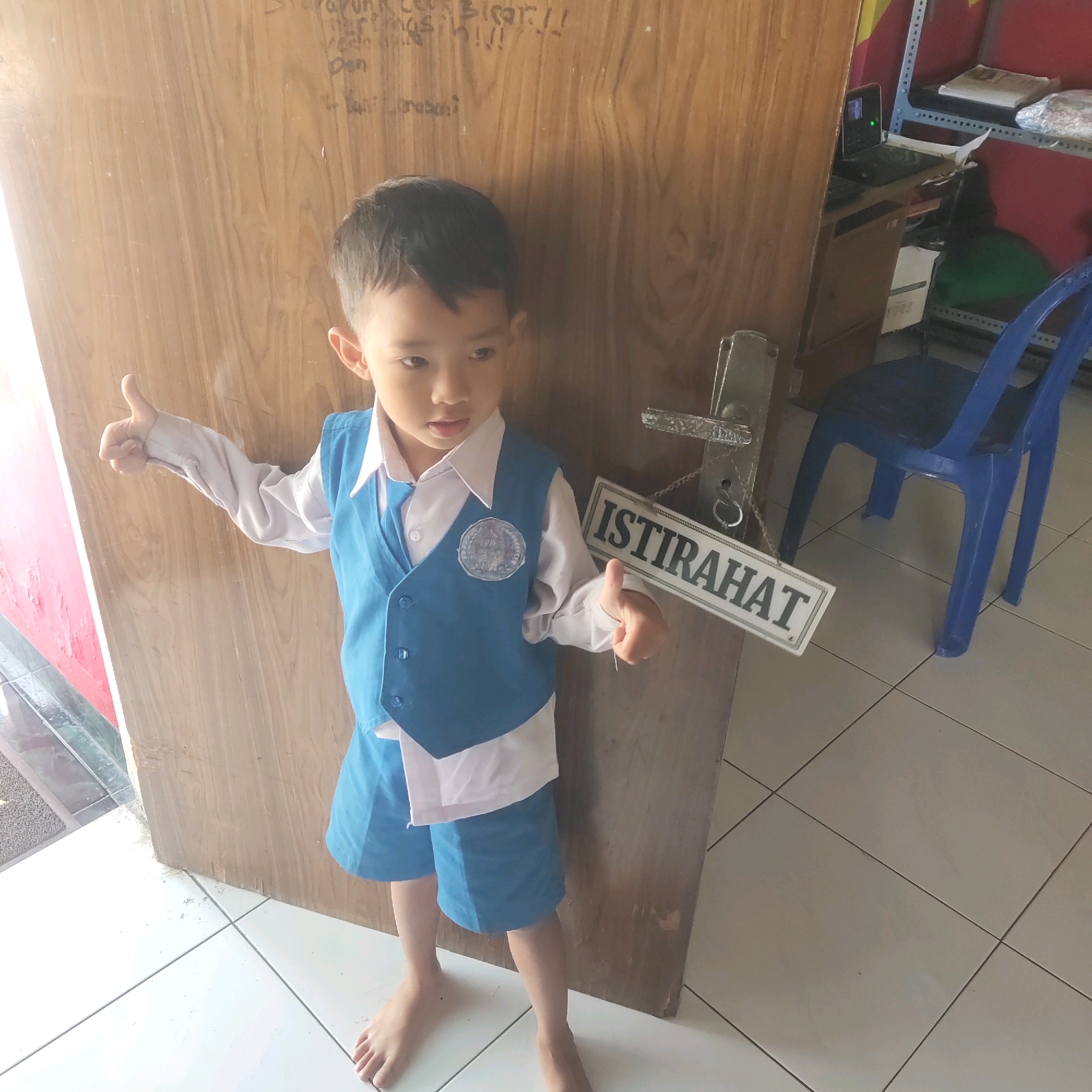 In Sragen, Indonesia, a young boy wearing a blue vest over a white shirt and blue shorts stands in front of a wooden door. He is giving a thumbs-up gesture with his left hand. A sign with the word "ISTIRAHAT" (which means "rest" in Indonesian) hangs from the door. To the right of the boy, a blue plastic chair is visible, and in the background, a shelf with items and a laptop on a desk can be seen. The floor is tiled. The lighting suggests it is daytime.