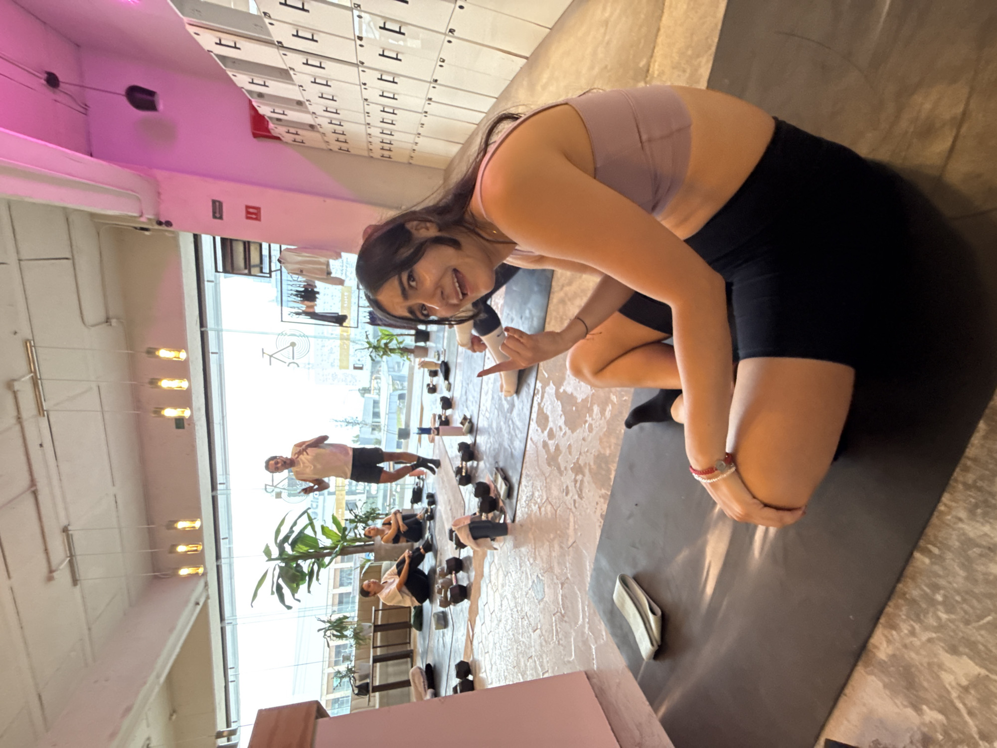 This image captures an indoor fitness studio scene in Puebla, Mexico, depicting a group workout session during the daytime.
The main subject is a young woman with dark hair, wearing a light purple sports bra and black athletic shorts. She is kneeling on a dark gray yoga mat, looking directly at the camera with a wide smile and making a hand gesture that resembles finger guns or pointing. On her right wrist, she wears a red and silver bracelet. A water bottle is visible near her.
In the background, several other individuals are engaged in exercises. One man in a light-colored t-shirt and dark shorts stands further back, while two women are seated on their yoga mats, surrounded by dumbbells. More dumbbells of varying sizes are scattered on the floor next to the mats, and a light gray resistance band rests on one mat.
The studio features a modern aesthetic with white walls, a high ceiling, and exposed pipes. A prominent feature is a wall of white lockers with numbers, where "11" or "1" is visibly repeated. Pink or purple accent lighting illuminates a beam along the ceiling. Large windows span one wall, allowing bright natural light into the space and offering a glimpse of the outside, though the view is indistinct. Several pendant lights hang from the ceiling, and potted plants add greenery to the environment. Clothing racks with apparel are also visible near the window. The floor has a textured, possibly concrete or patterned, finish.
The overall atmosphere suggests an energetic and friendly workout environment.