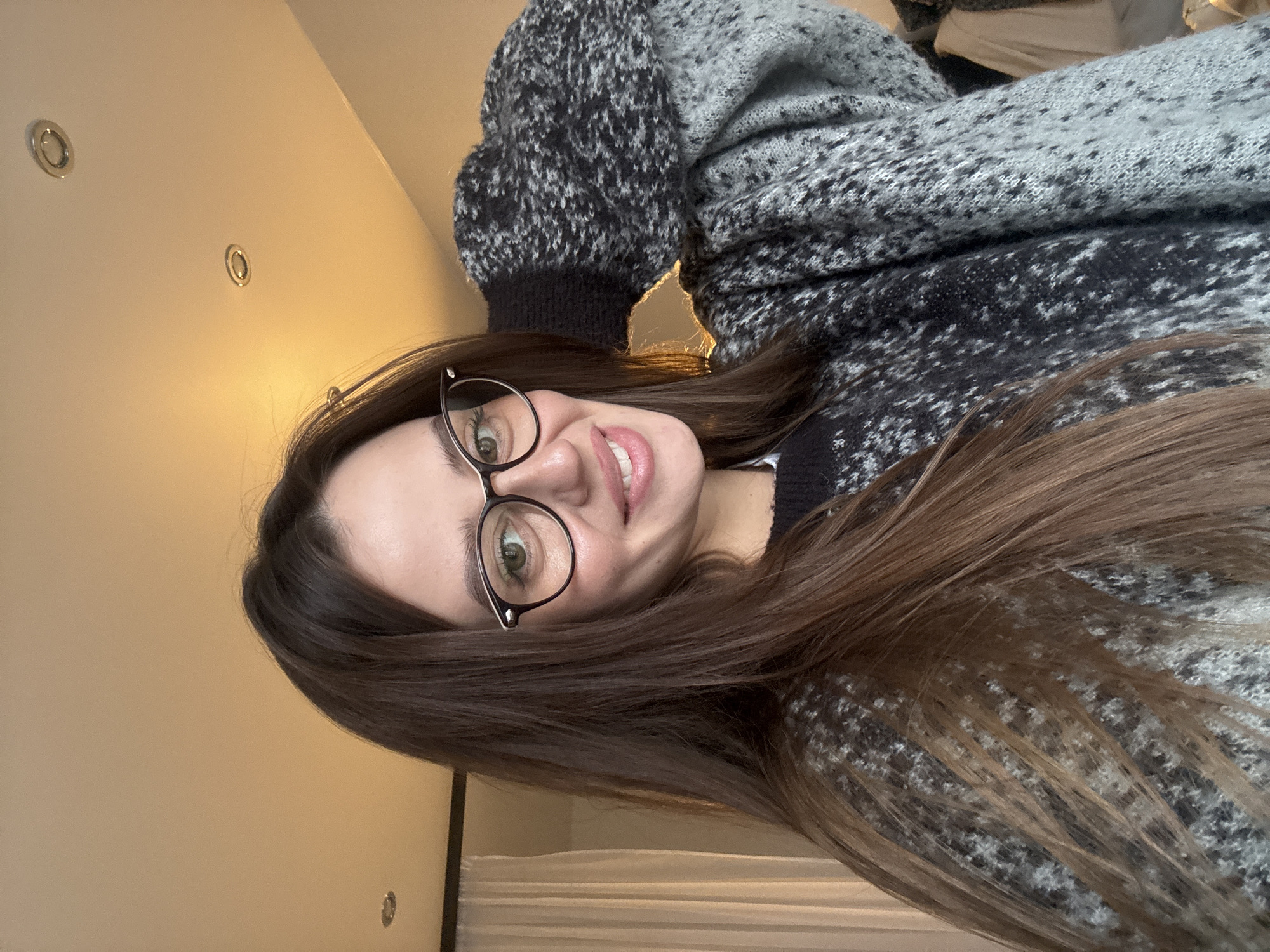 A young woman with long brown hair and glasses is facing the camera and smiling. She is wearing a black and gray patterned sweater. The background is a light beige wall with recessed lighting. The location in Krasnoyarsk, Russia, cannot be determined from this image.