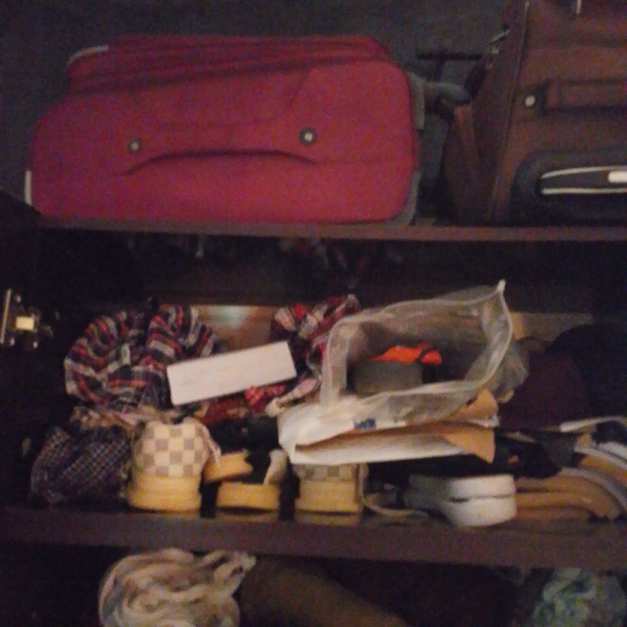 The image shows a shelf filled with various items, likely in a storage area. On the top shelf, a large red suitcase occupies the left side, with a smaller brown suitcase to its right. The red suitcase has a visible white stripe on its side and a circular emblem or handle. The brown suitcase also has white detailing.
The middle shelf is cluttered with a variety of objects. To the left, a checkered fabric bundle sits next to a dark patterned bag. In the center, a white plastic bag contains several items, including orange-colored objects. There's also a white rectangular piece of paper or cardboard on this shelf. Further to the right, two round objects, one yellow with a checkered pattern and another yellow, are visible.
The bottom shelf displays more items, including a light-colored, possibly fabric bundle with a swirling pattern on the far left. To the right, white objects resembling shoes or shoe soles are visible. The overall environment appears to be a dimly lit indoor space with shelves constructed from a dark material. The image does not provide any information or visual cues that would allow for identification of its location in Jalingo, Nigeria.